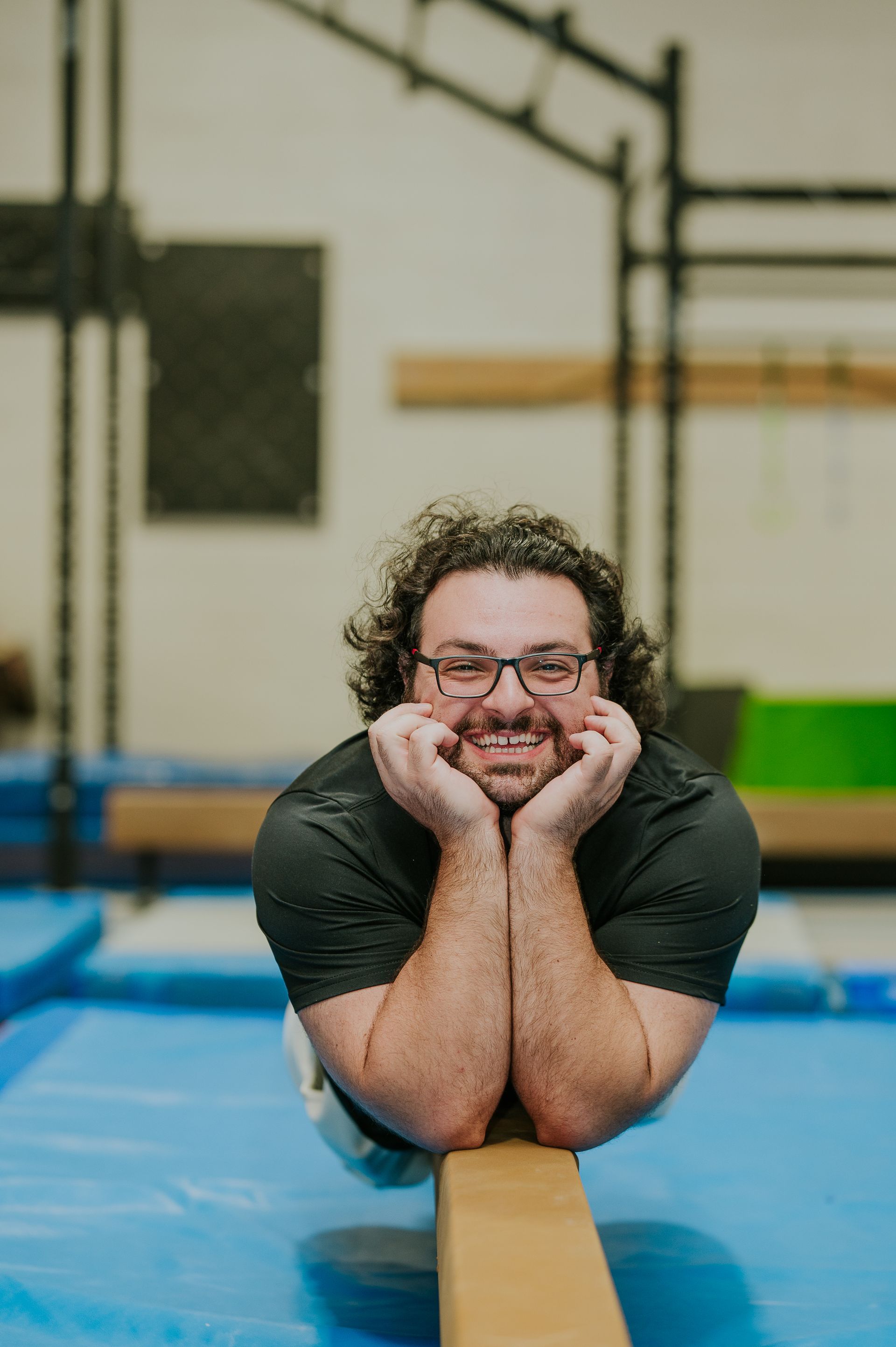 Man balancing on a beam in a gym, leaning on his hands and smiling