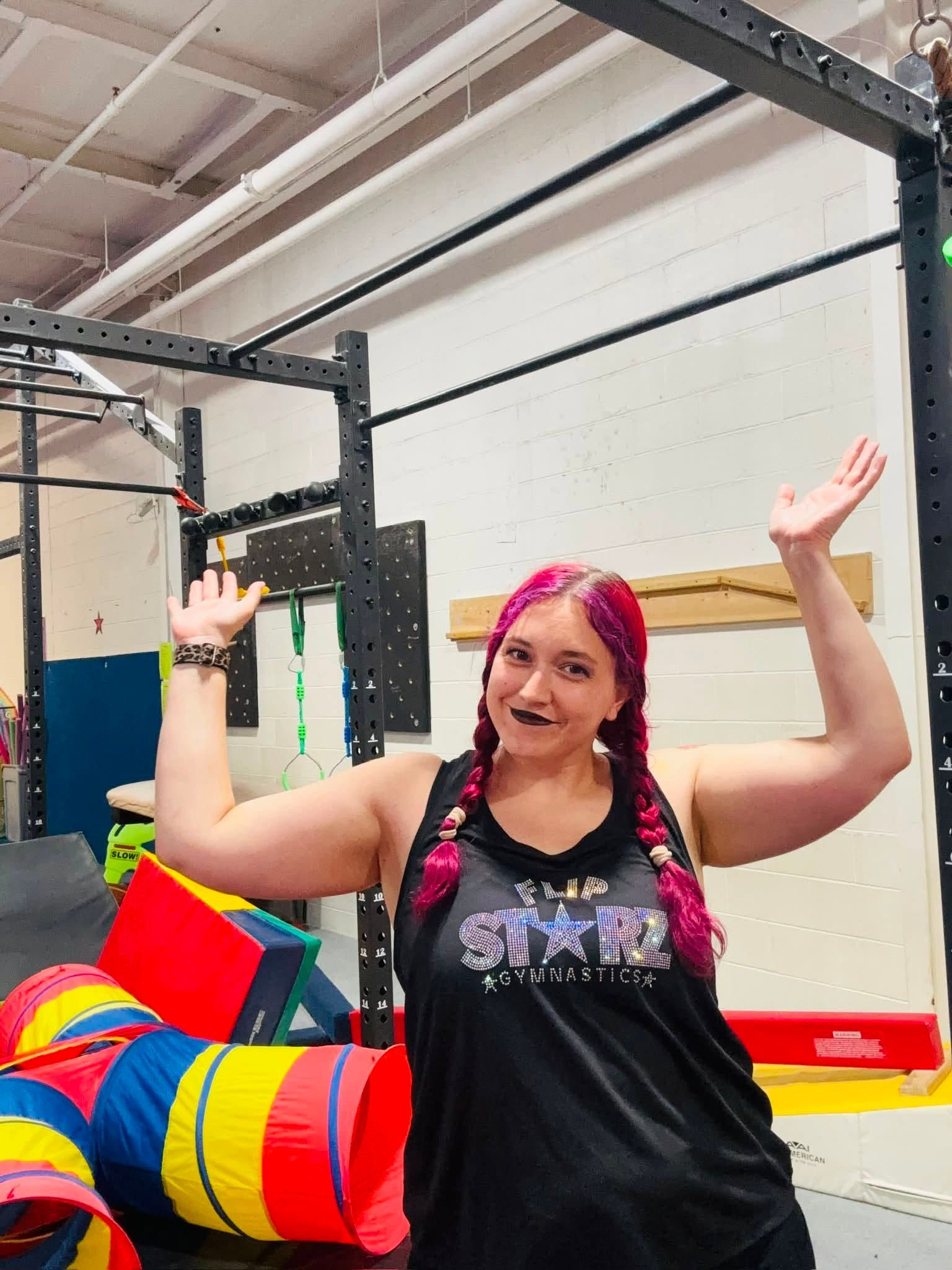 Woman with pink hair smiling and waving in a gym, beside colorful mats and obstacle bars.