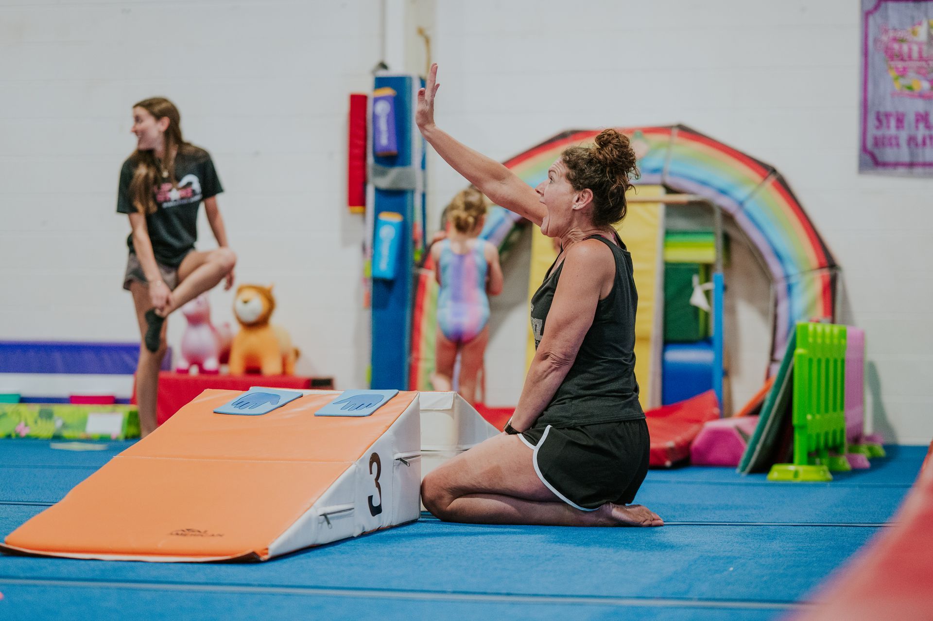 Woman kneeling on blue mat in gym, reaching for a pole beside an orange ramp and rainbow arch