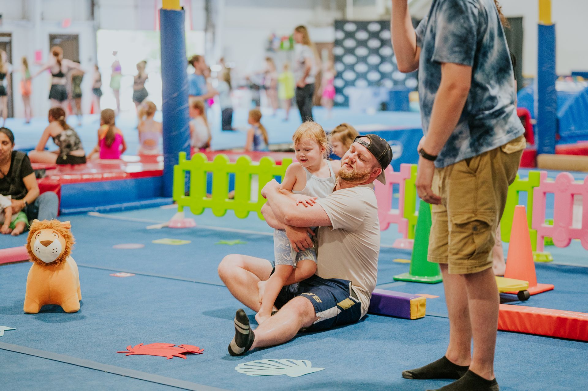 Adult kneeling and holding a smiling child in a colorful indoor play area with other people nearby
