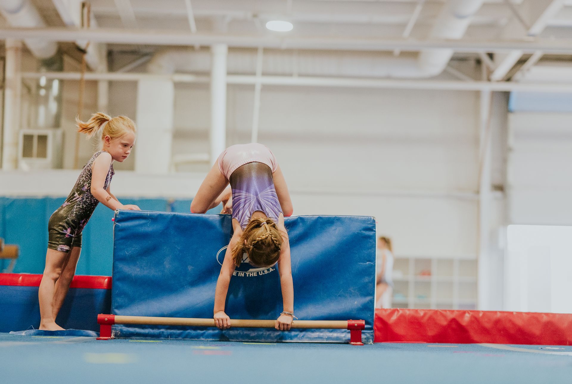 Two children on a gymnastics mat, one holding a blue block while the other hangs upside down over it