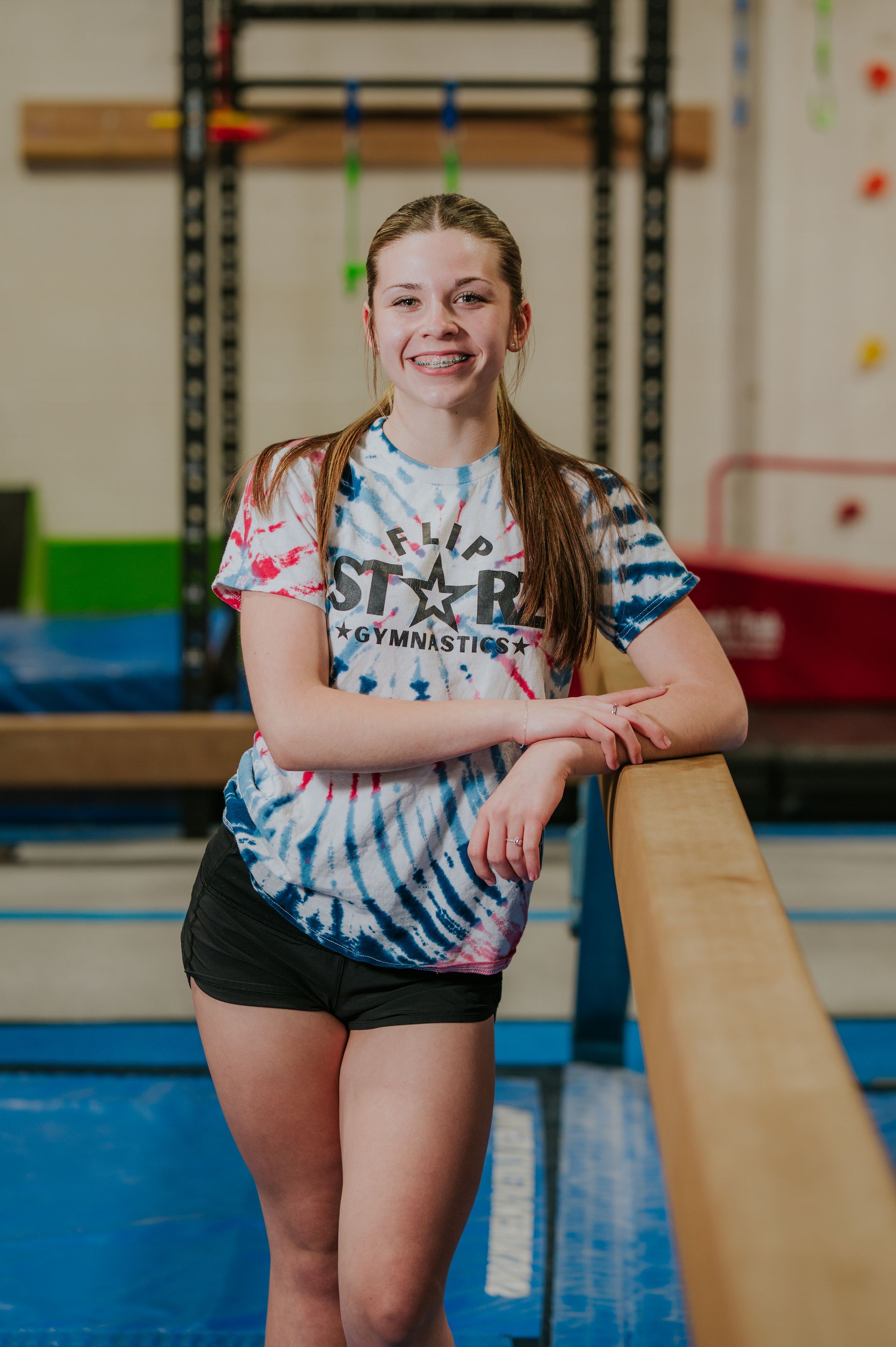 Smiling gymnast leaning on a balance beam in a gym, wearing a tie-dye shirt and black shorts