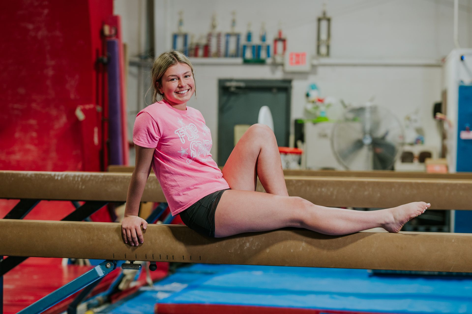 Gymnast in pink shirt sitting on a balance beam in a gym, smiling