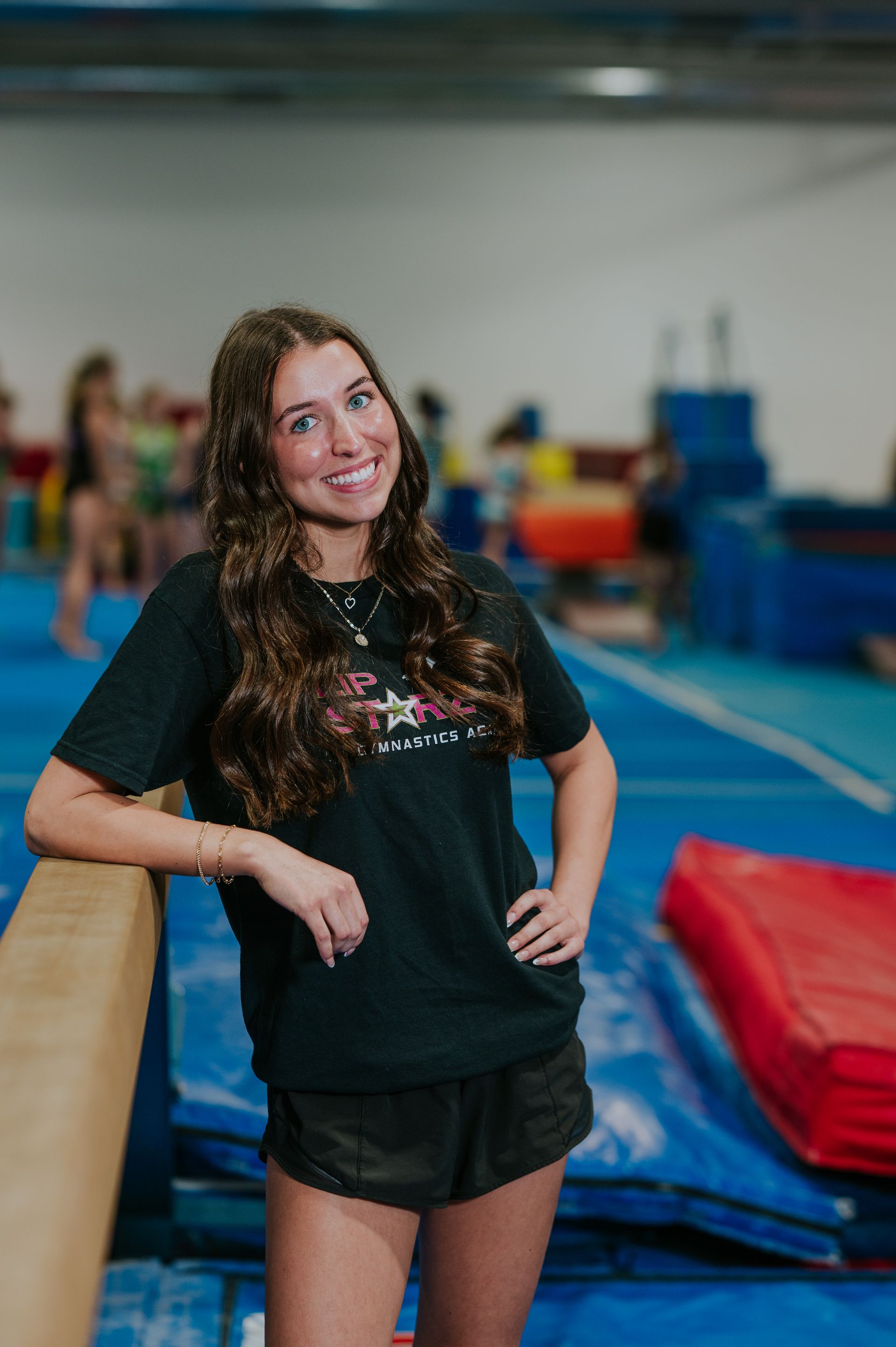 Woman smiling beside a balance beam in a gym, with mats and equipment in the background
