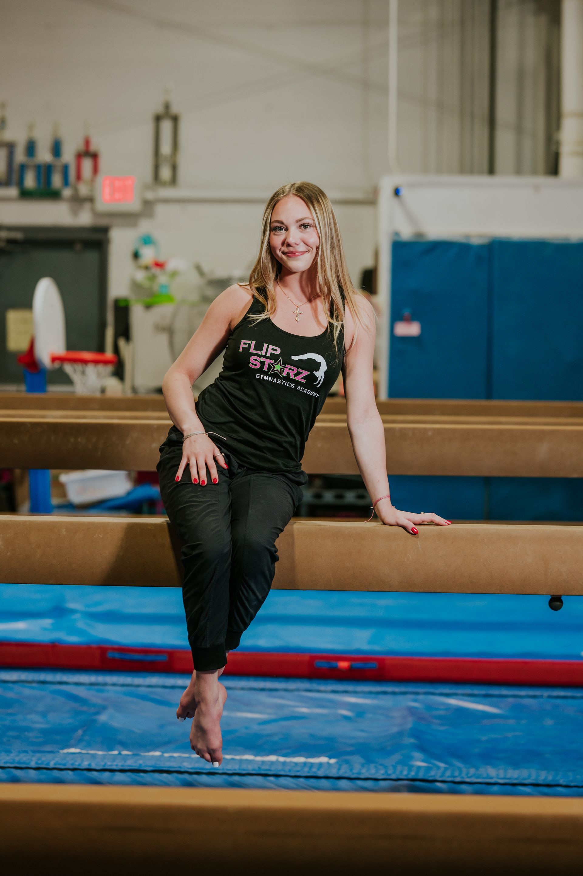 Woman in a gym leaning on a balance beam, smiling in a black tank top and leggings.