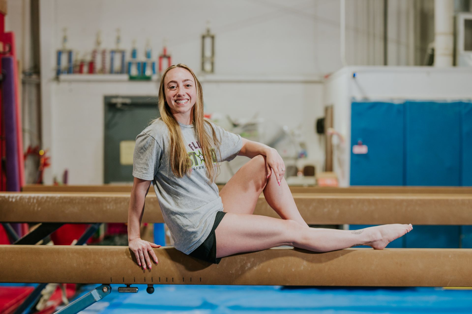 Gymnast balancing on a beam in a bright training gym, smiling and posing with one leg extended.