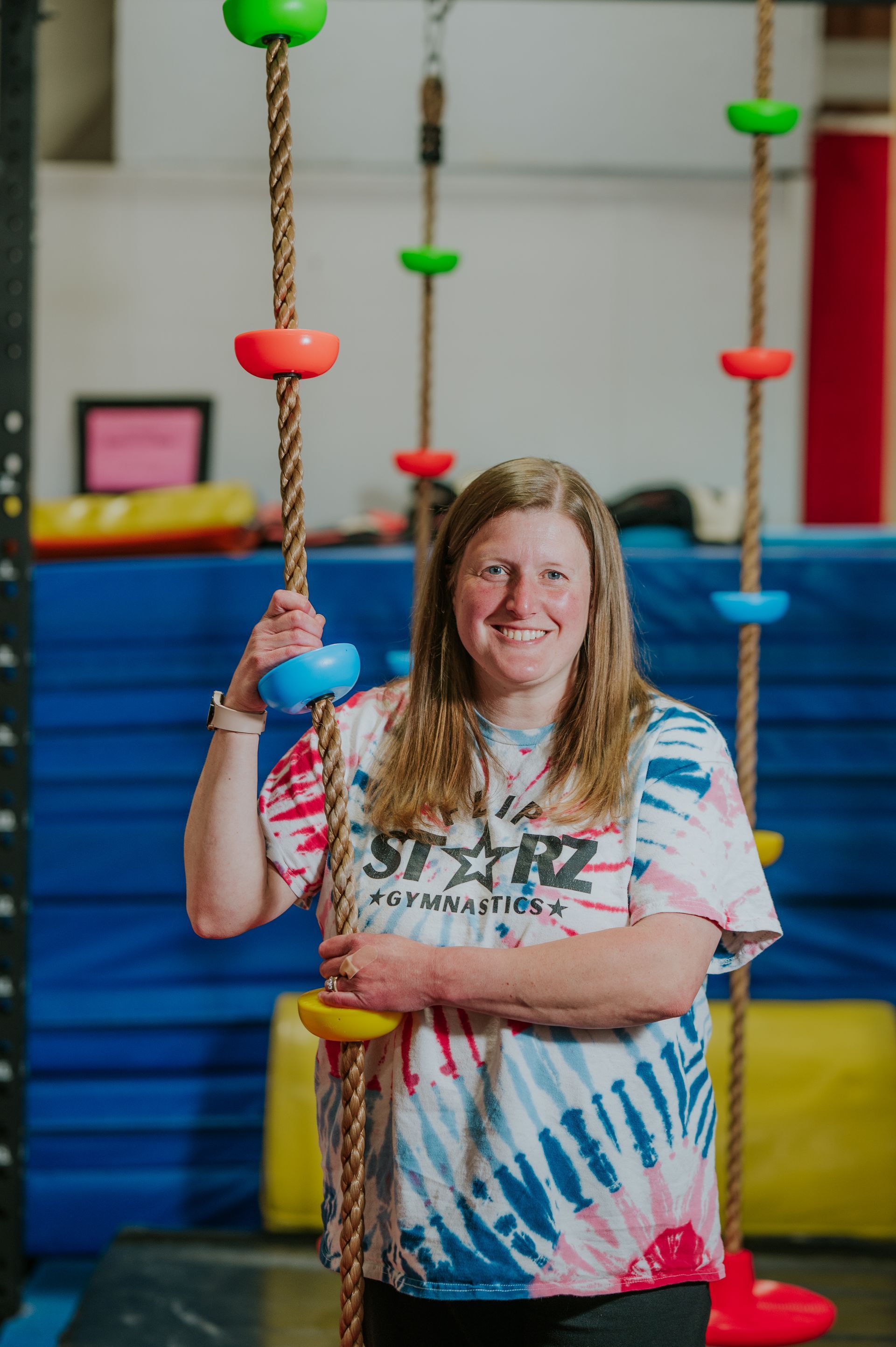 Person smiling and holding a climbing rope in an indoor play area with colorful ropes and blue steps