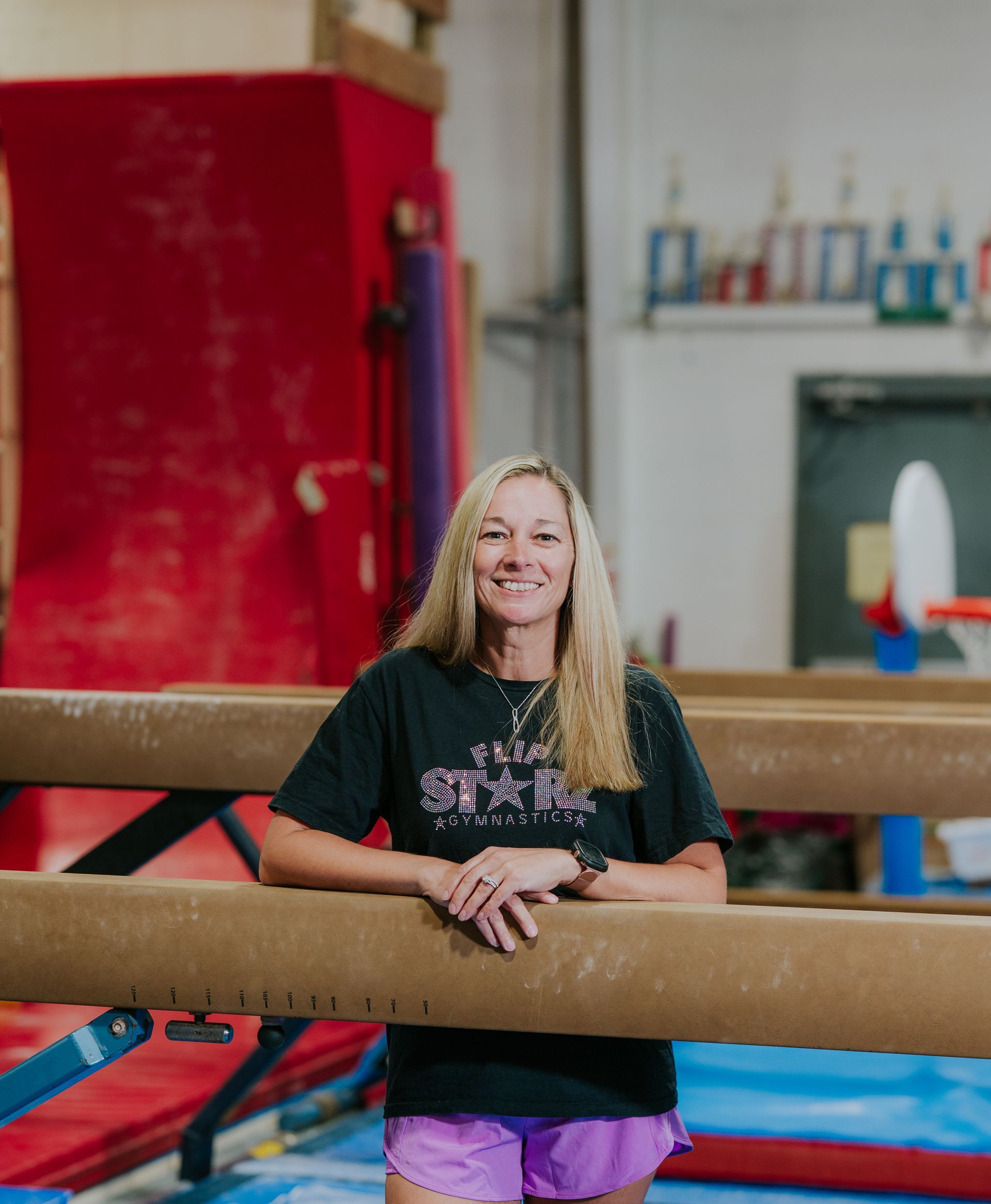Woman leaning on balance beams in a gym, smiling at the camera.