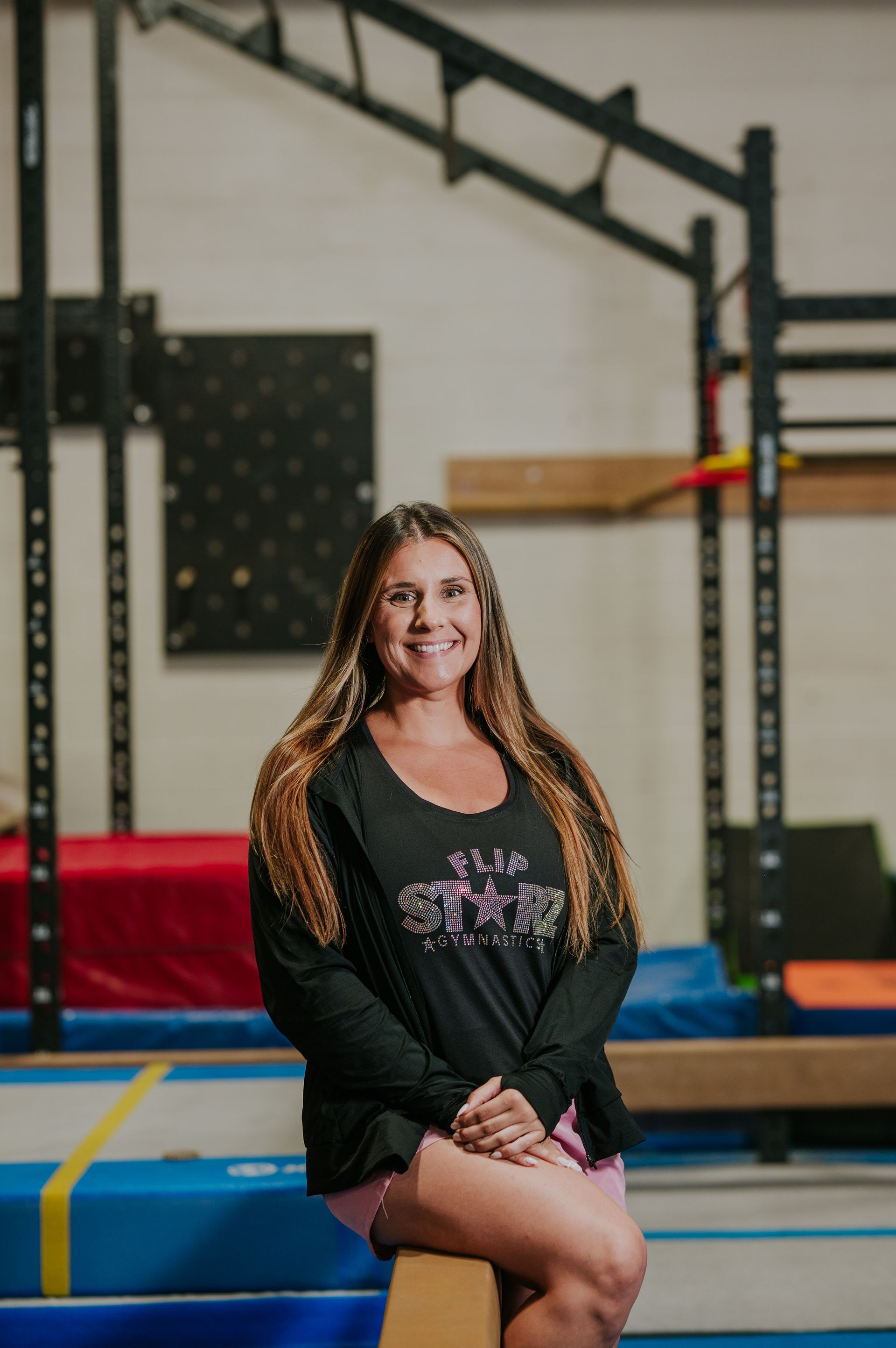 Woman seated on a blue gym mat in a workout space, wearing a black top and shorts, smiling at the camera.