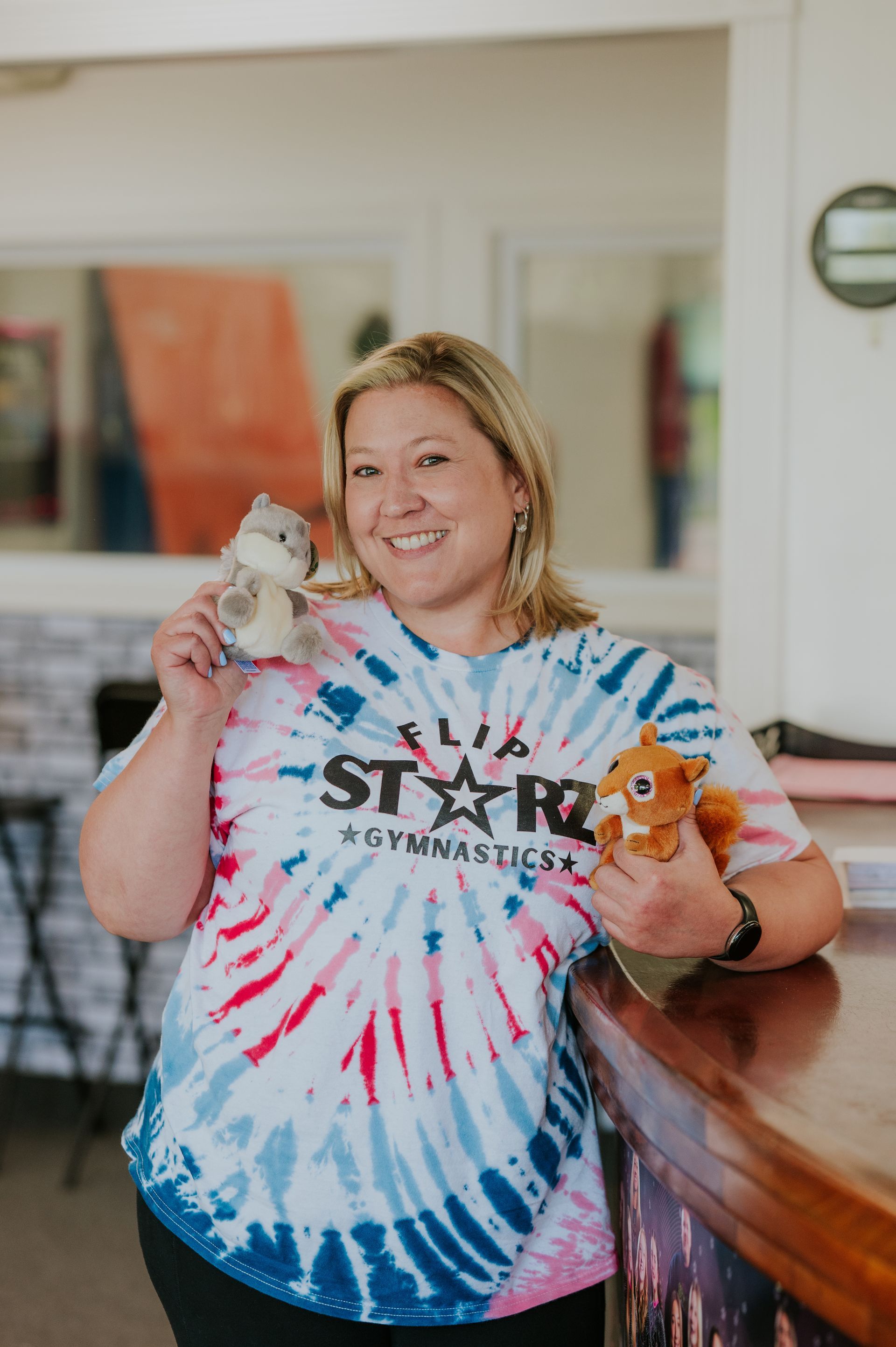 Person in tie-dye shirt holding two small animals and smiling in an indoor setting
