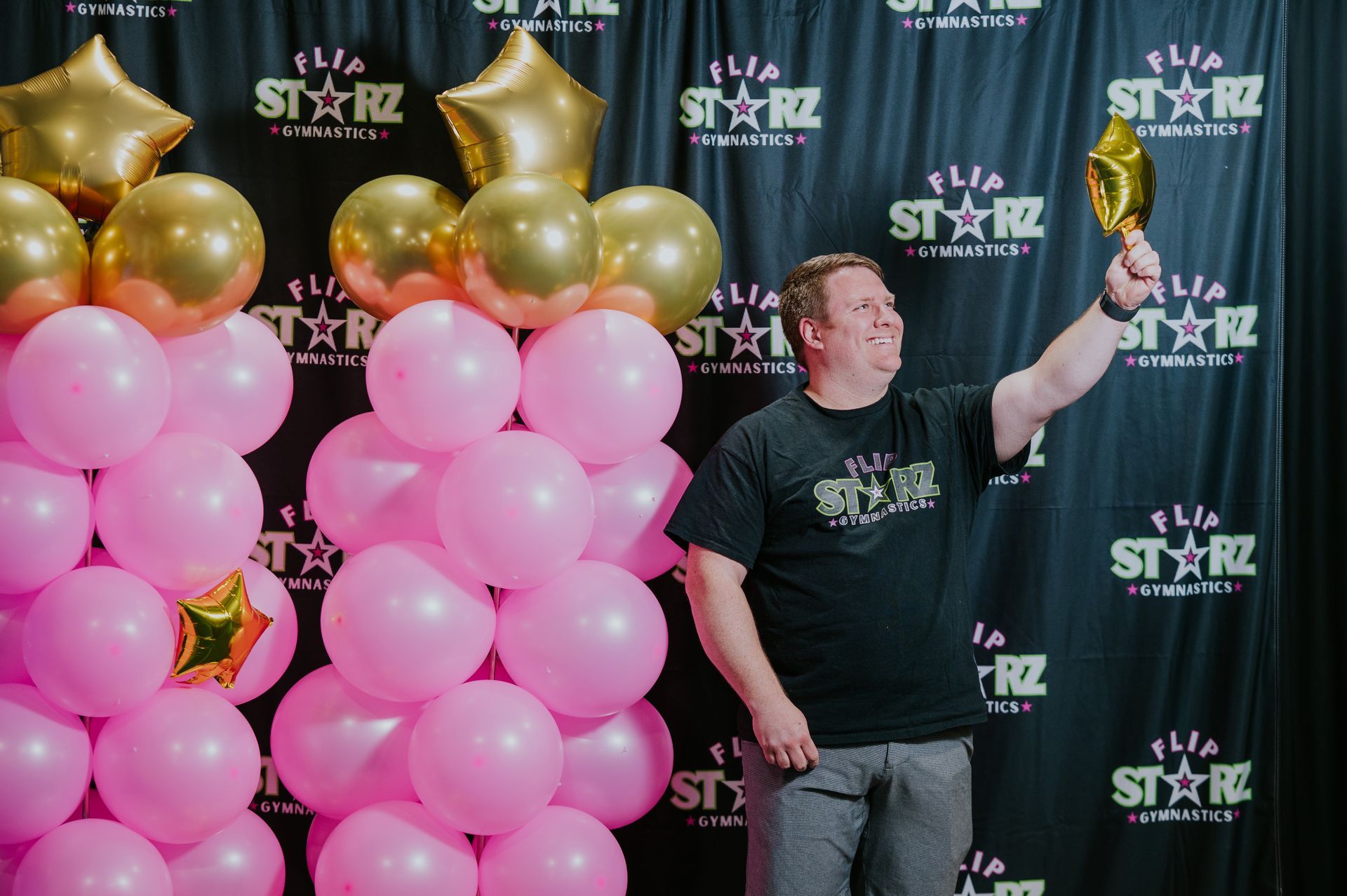 Man posing with a raised trophy beside pink and gold balloon columns at a STAR backdrop