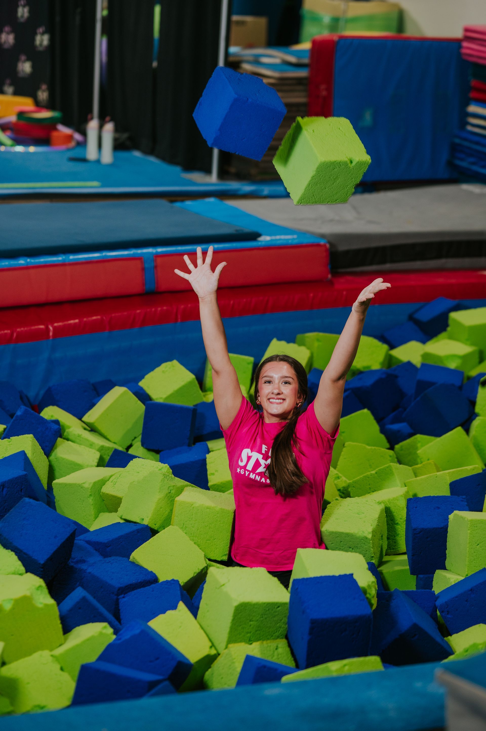 Person in a pink shirt with arms raised in a foam pit at an indoor gym
