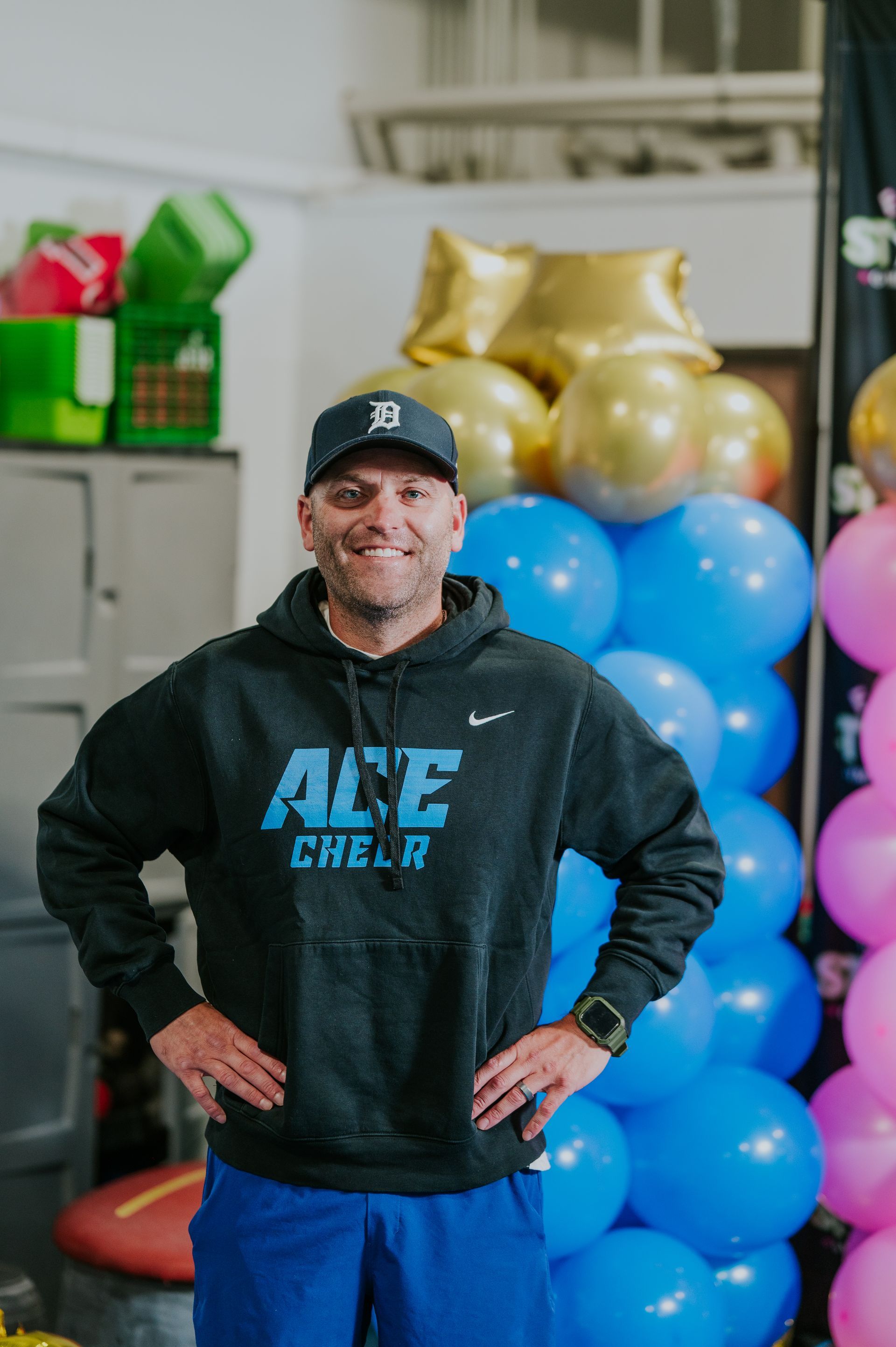 Man in black hoodie and cap stands with hands on hips beside stacked blue exercise balls in a gym.