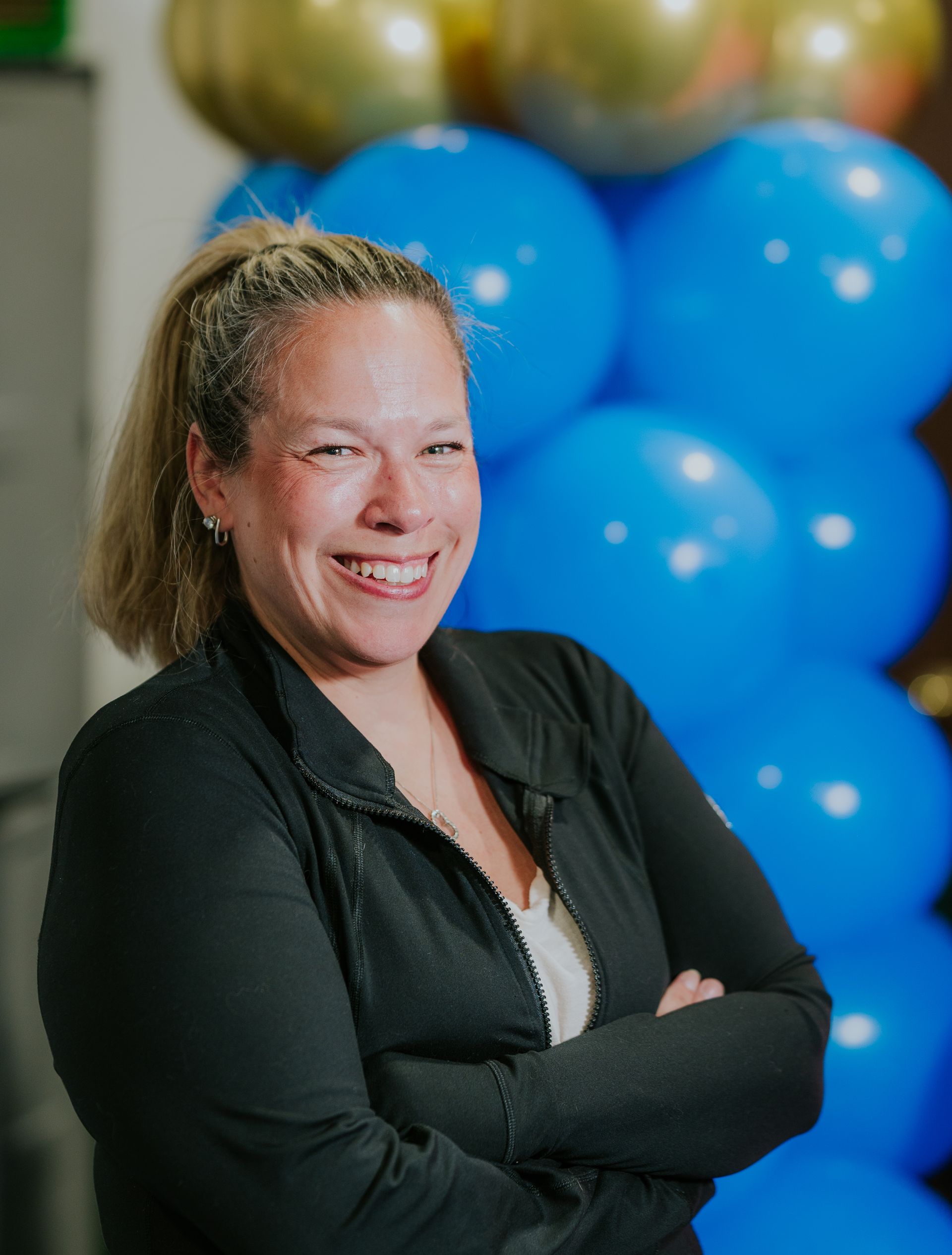Smiling person with arms crossed in front of blue and gold balloons
