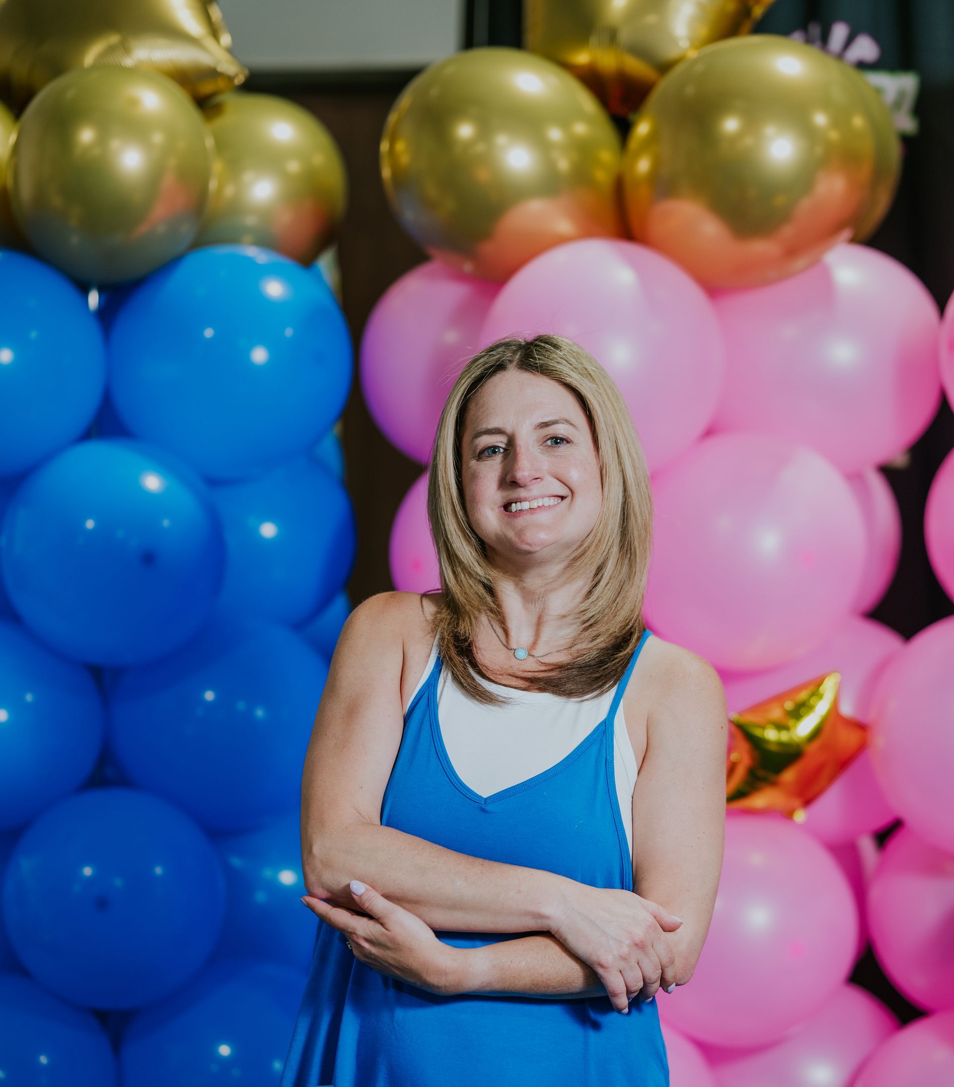 Woman in blue top smiling with arms crossed in front of blue, pink, and gold balloons