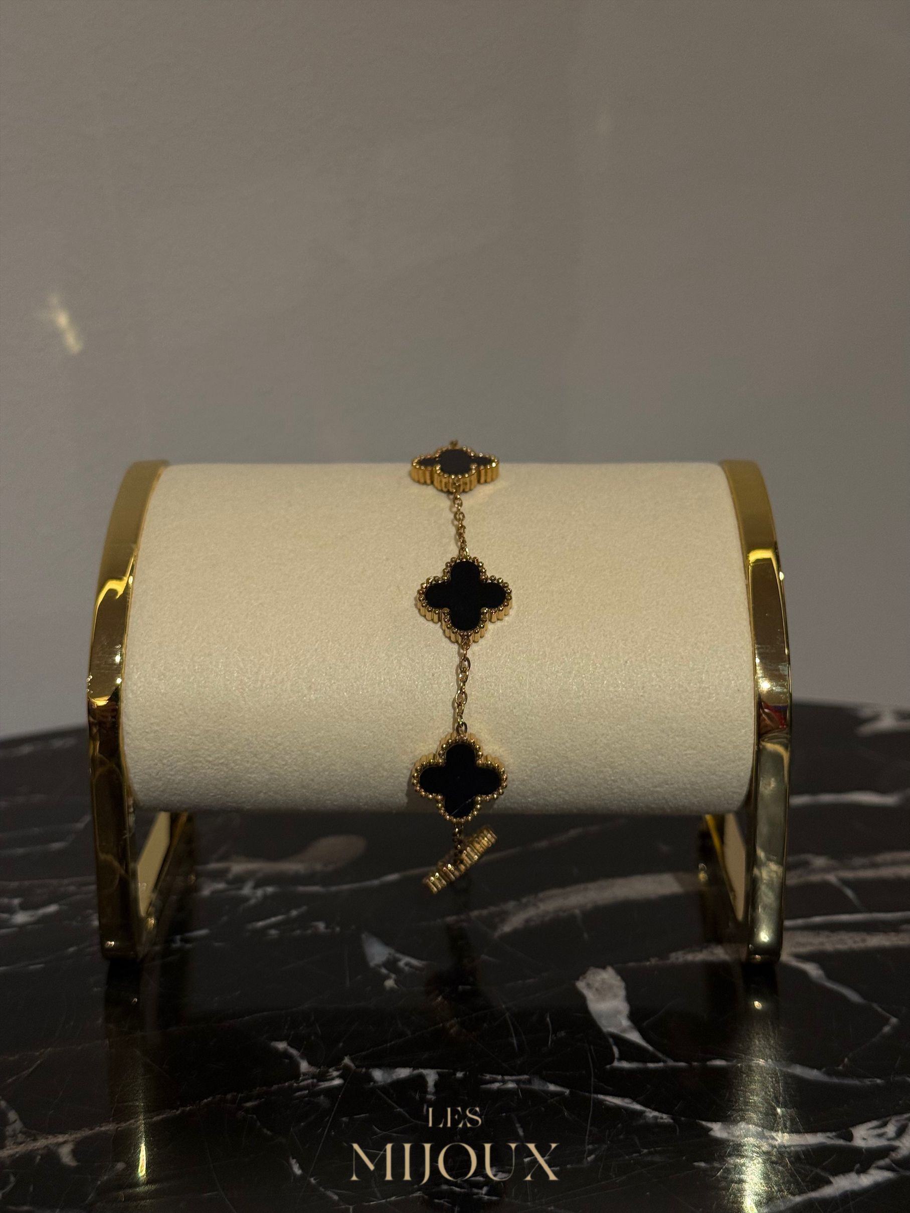 Gouden armband met drie zwarte klavermotieven op een displaystandaard, tegen een marmeren ondergrond.