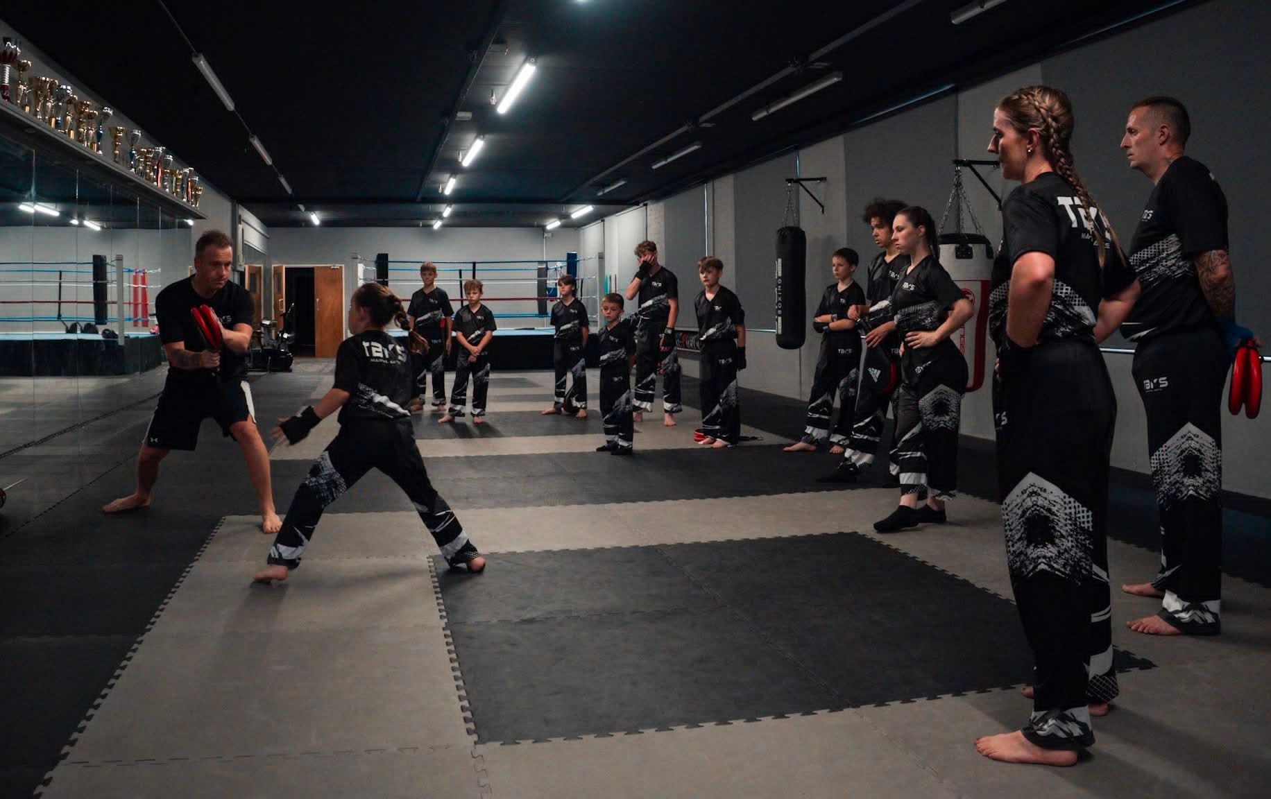 A coach talks to a young martial artist in a ring; the child wears gear, preparing to compete.