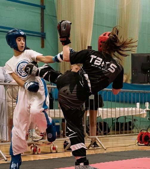 Two kickboxers in a match. One in white, the other in black, kicking. Indoor setting.
