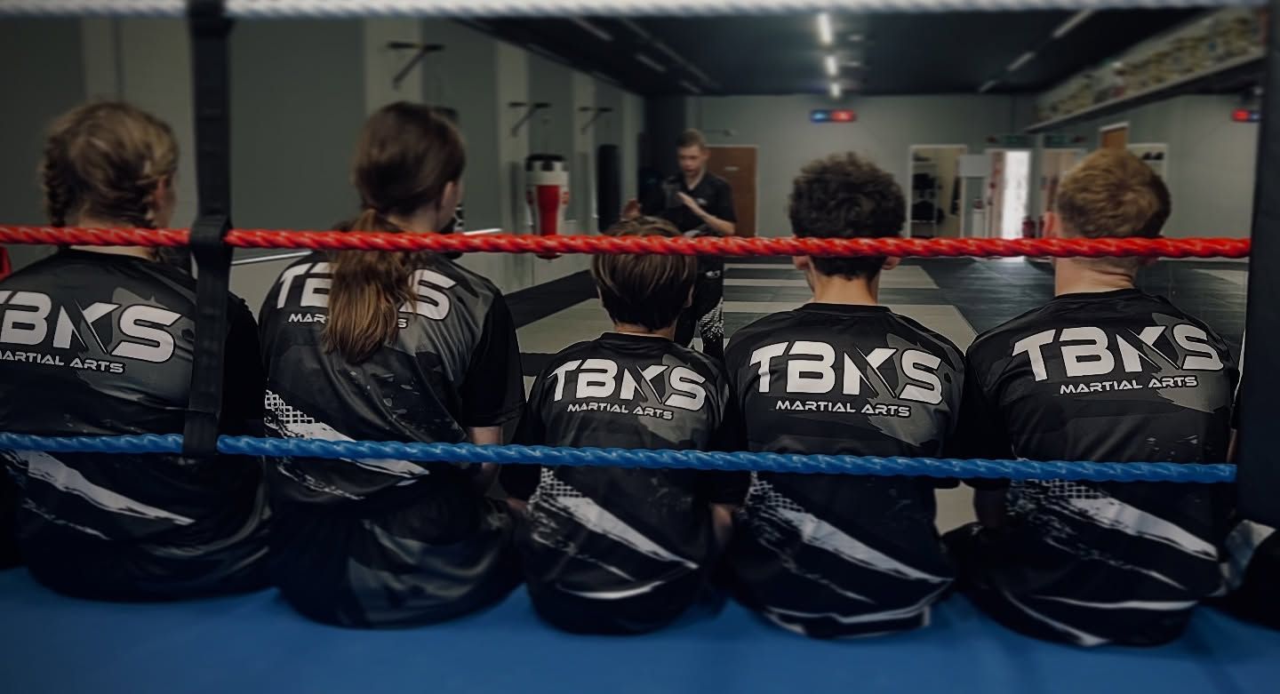 Five children in black jackets watch a martial arts instructor in a boxing ring.