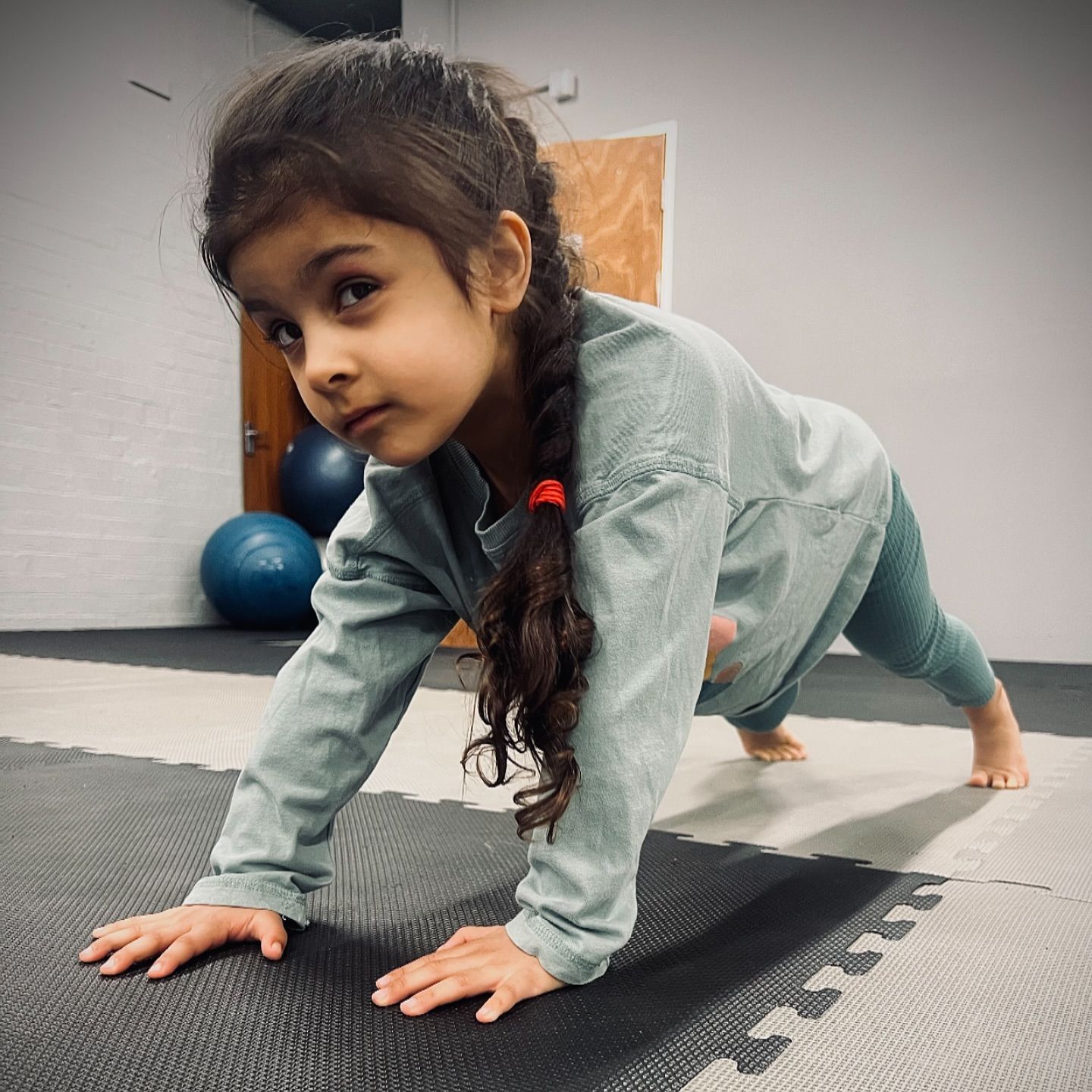 A young girl with a braid doing a push-up on a mat, looking at the camera.