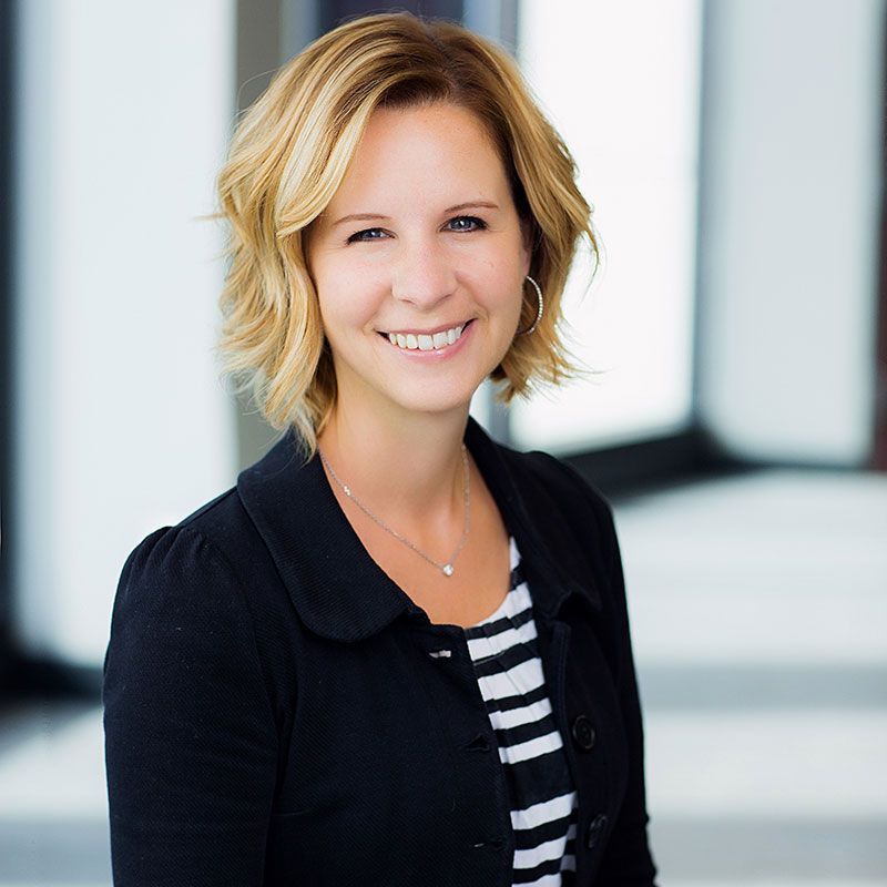 A smiling person with shoulder-length blonde hair, wearing a black blazer over a black-and-white striped shirt.