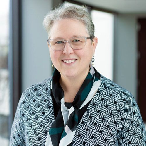 Smiling person with short gray hair and glasses, wearing a geometric-patterned shirt and a dark green scarf indoors.