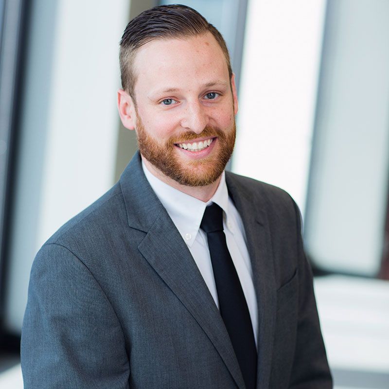Professional headshot of a person with a beard, wearing a gray suit, white shirt, and black tie, smiling at the camera.