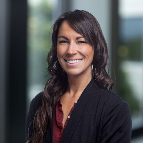 A professional headshot of a smiling person with shoulder-length dark hair, wearing a dark blazer over a maroon shirt.