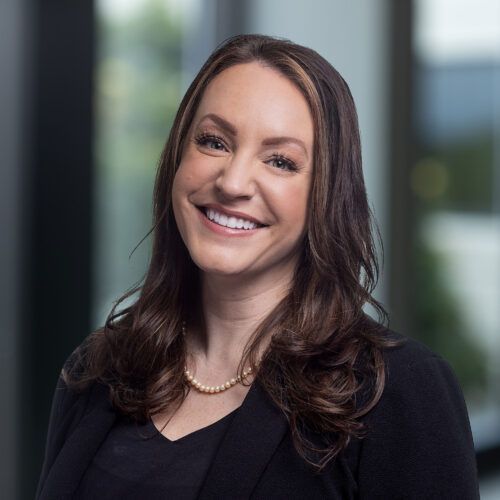 A smiling professional in a black blazer and pearl necklace, set against a blurred office background.