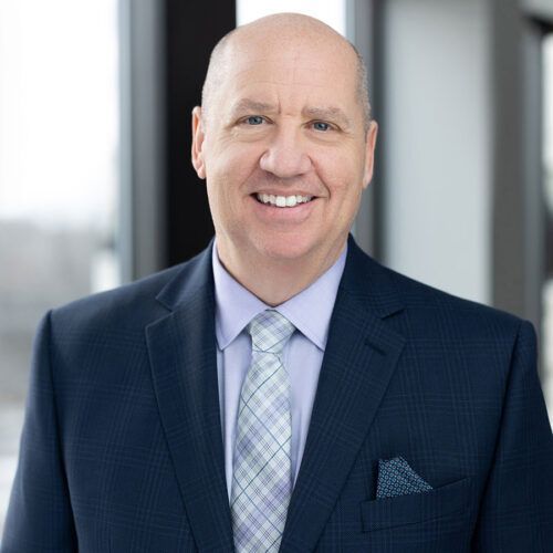 A person in a dark blue suit, light shirt, and patterned tie smiling for a professional headshot in an office setting.