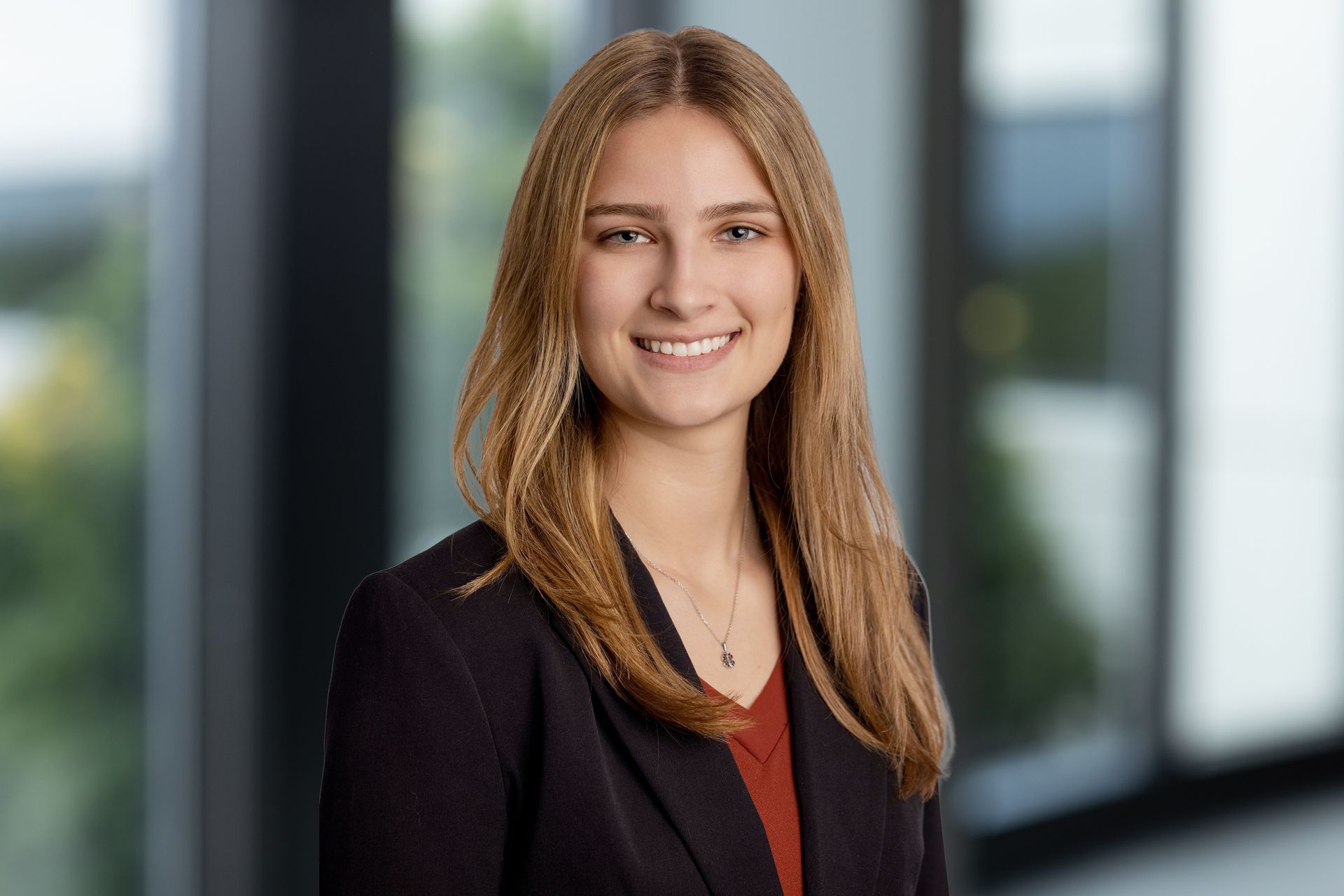 A professional headshot of a person with long light brown hair smiling at the camera, wearing a blazer in a modern office.