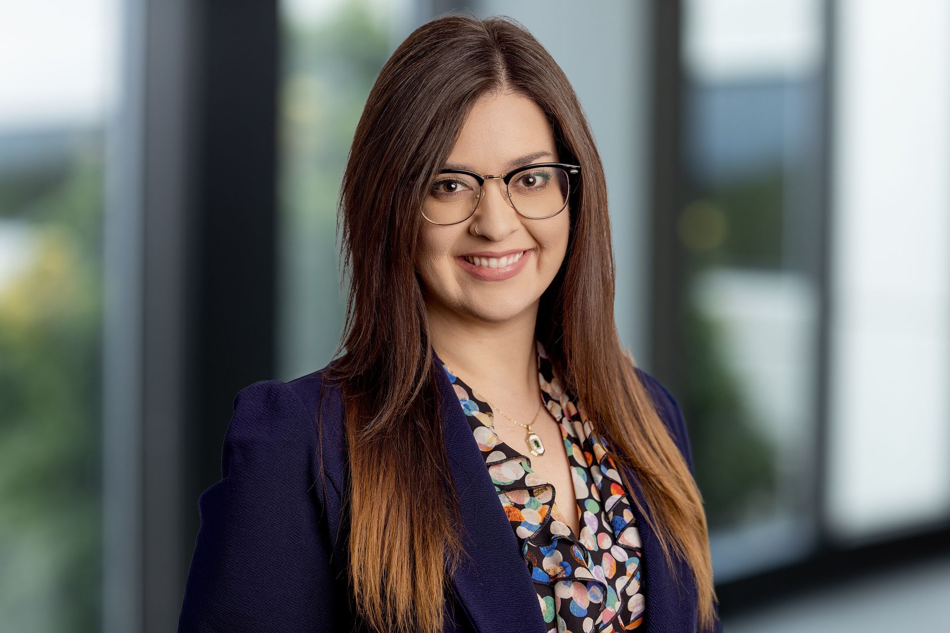 A smiling professional with glasses wears a tan blazer over a floral green top and a gold pendant necklace.