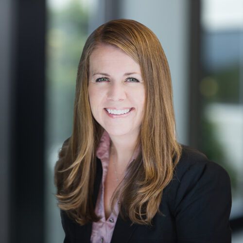 Professional headshot of a smiling person with long brown hair, wearing a light-colored blouse and a dark blazer.