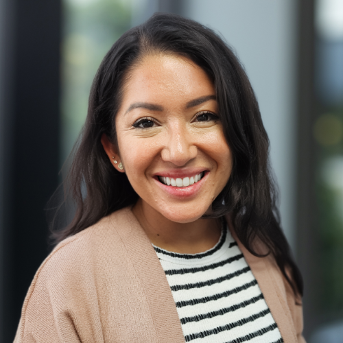 Smiling woman in a beige cardigan and striped shirt, standing outdoors with a blurred background