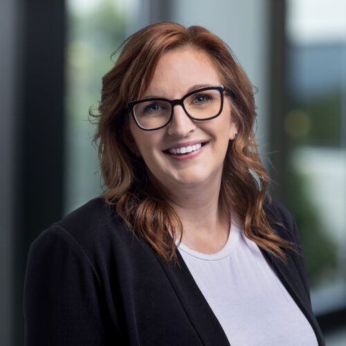 A person with shoulder-length auburn hair and black glasses, smiling in a black blazer over a white shirt, against a blur.