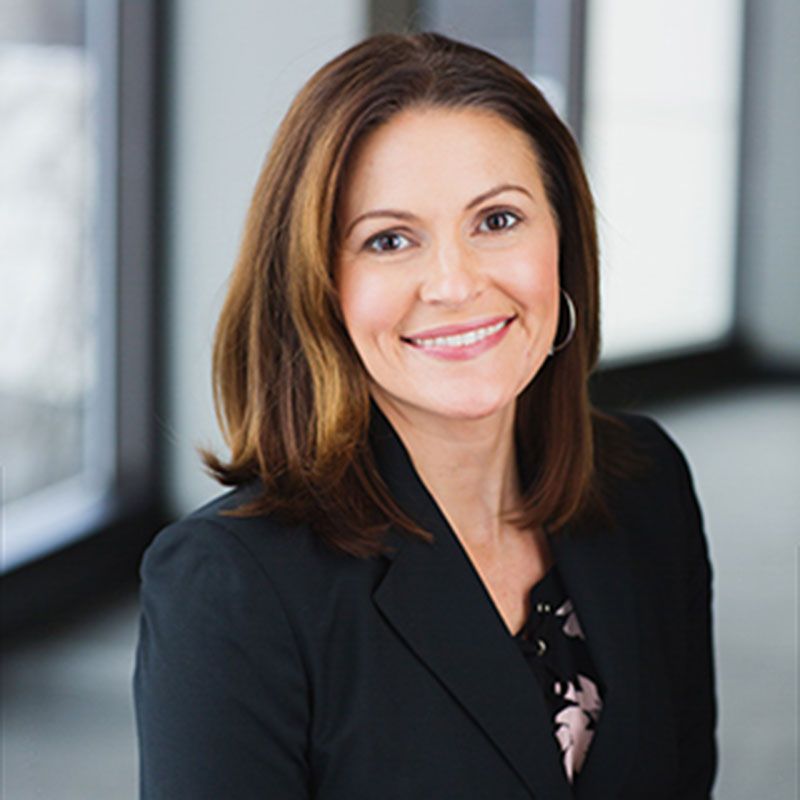 A smiling professional with shoulder-length brown hair, wearing a black blazer over a patterned top in a bright office.