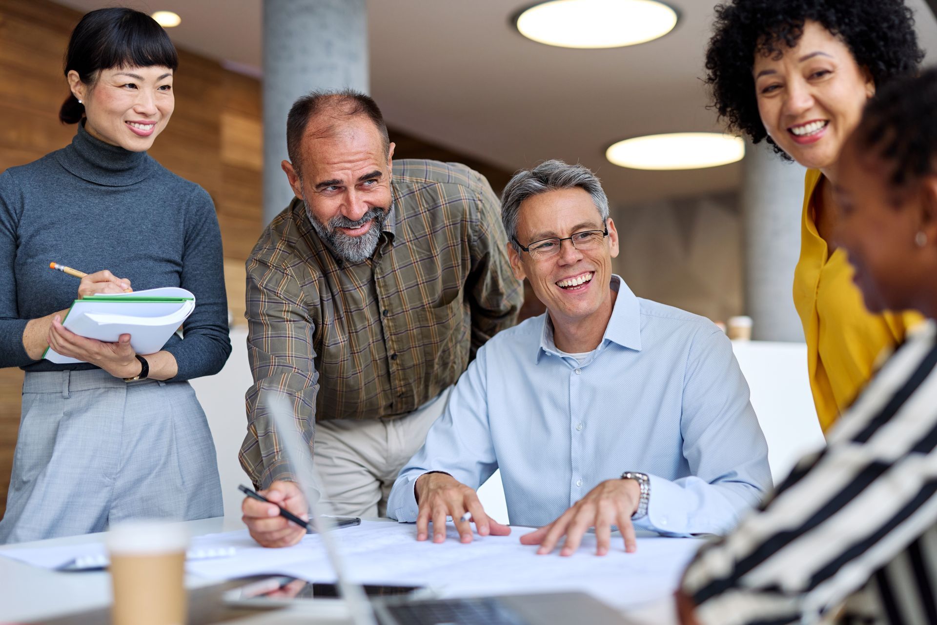 A group of professionals in a bright office standing around a desk, collaborating on documents and smiling.