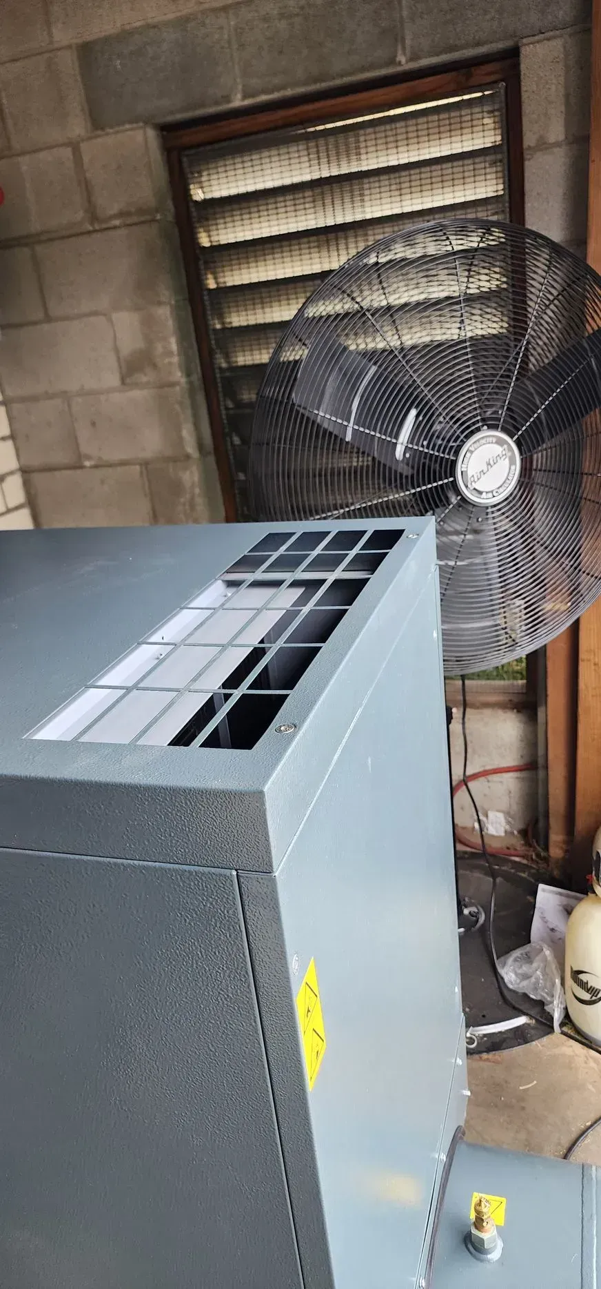 A large gray metal box with a vent, a fan, and a louvered window in a cinder block room.