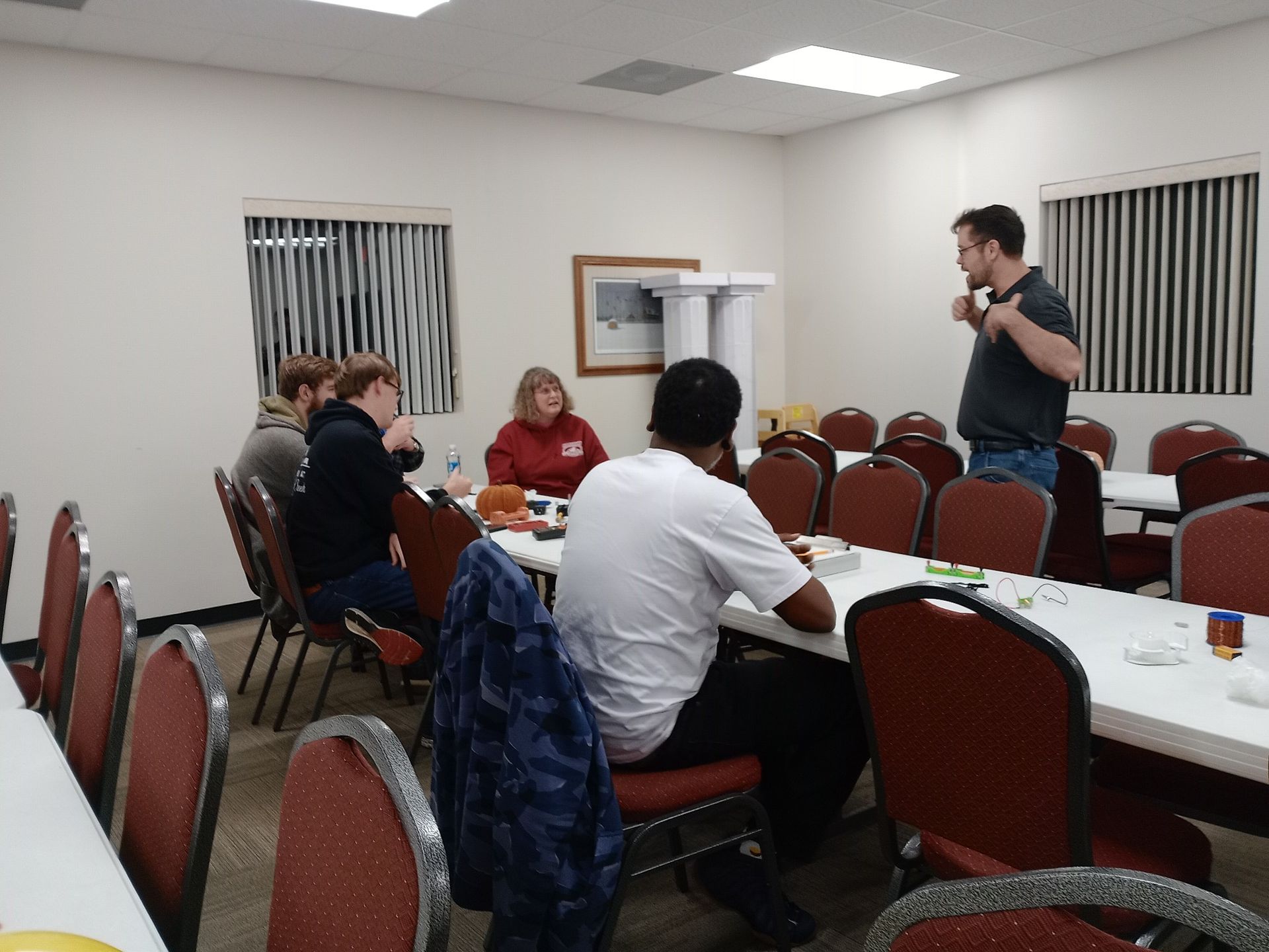 People seated at tables in a room, some talking, one person standing and speaking, likely a meeting or event.