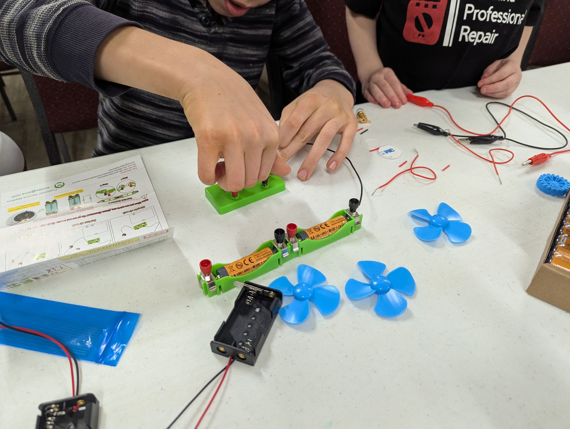 Two children assembling a circuit with wires, battery, and fan blades on a white table.