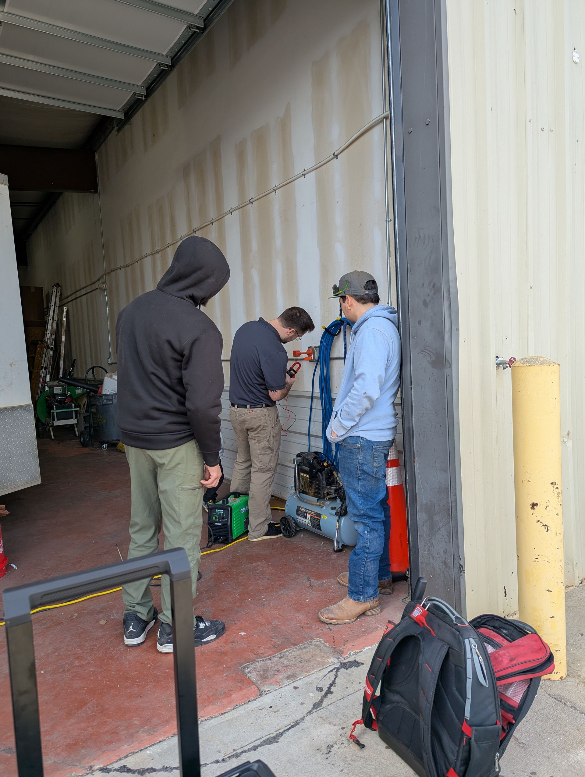 Three people working near equipment by a loading dock. One in a hoodie, one in a blue shirt, and one in a light blue jacket.