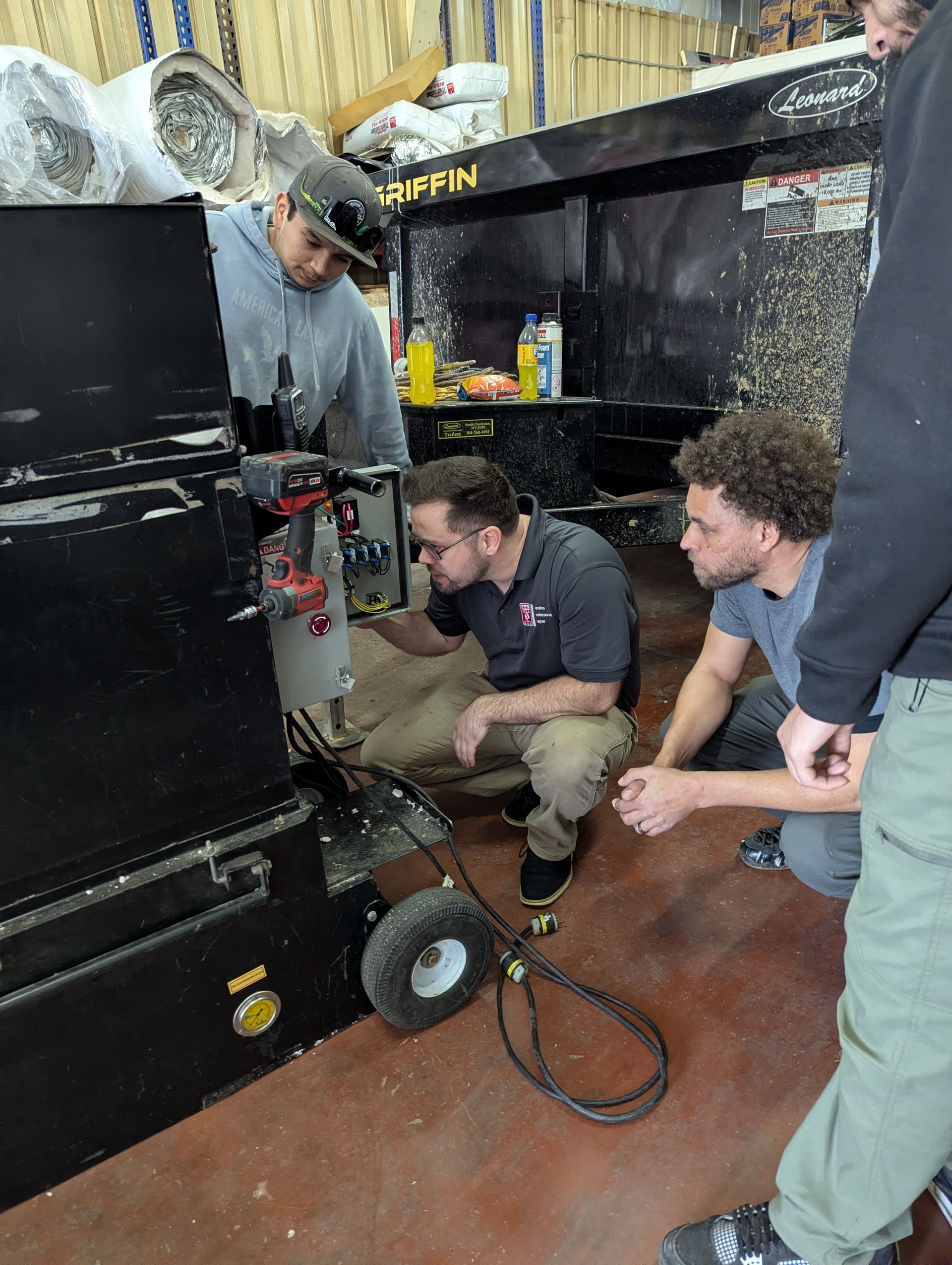 Four people examining machinery in a workshop. One kneels, pointing; others observe. Dark equipment, tools visible.