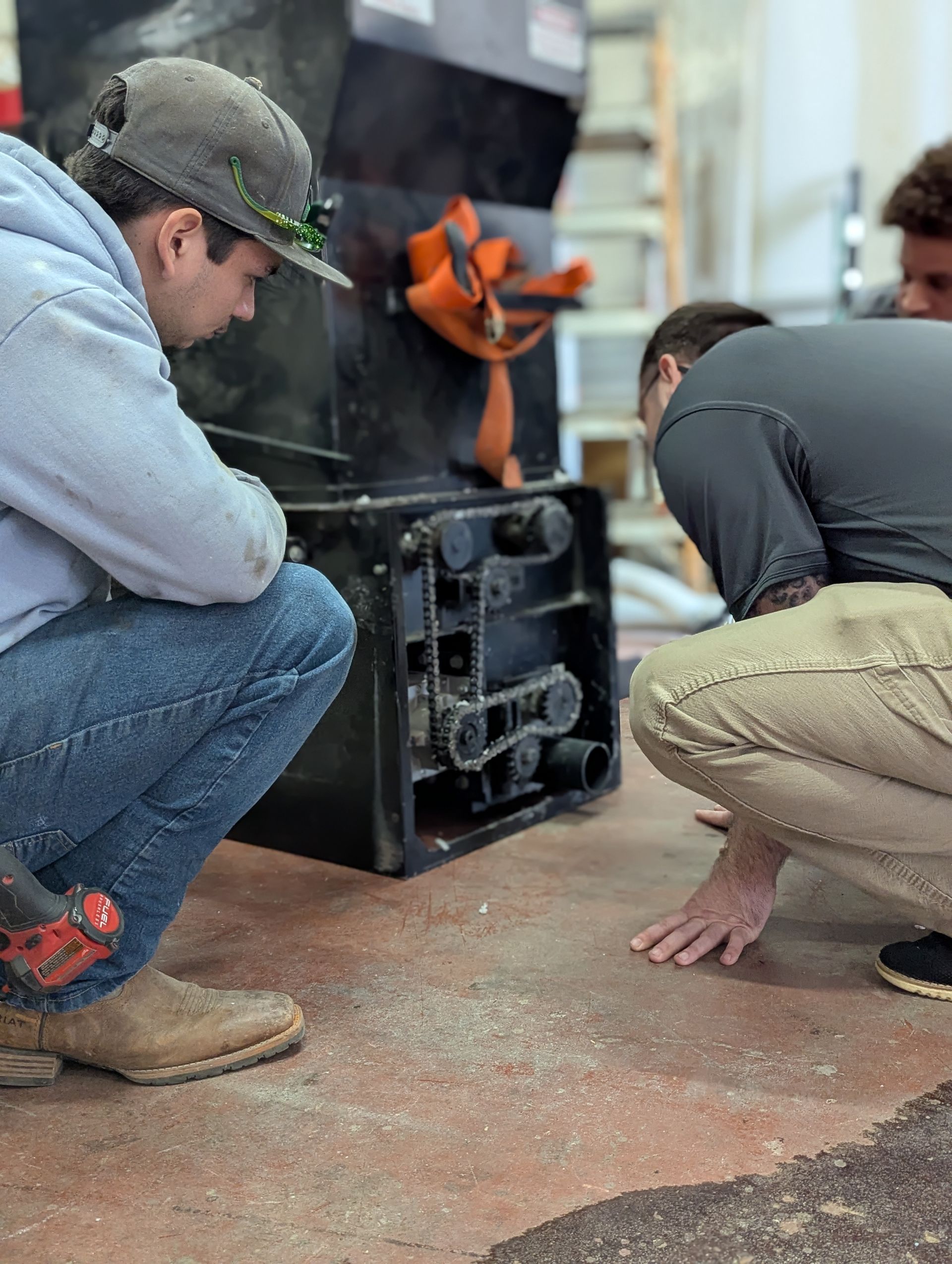 Three people examining a black machine in a workshop. One squats with a tool, two lean in.