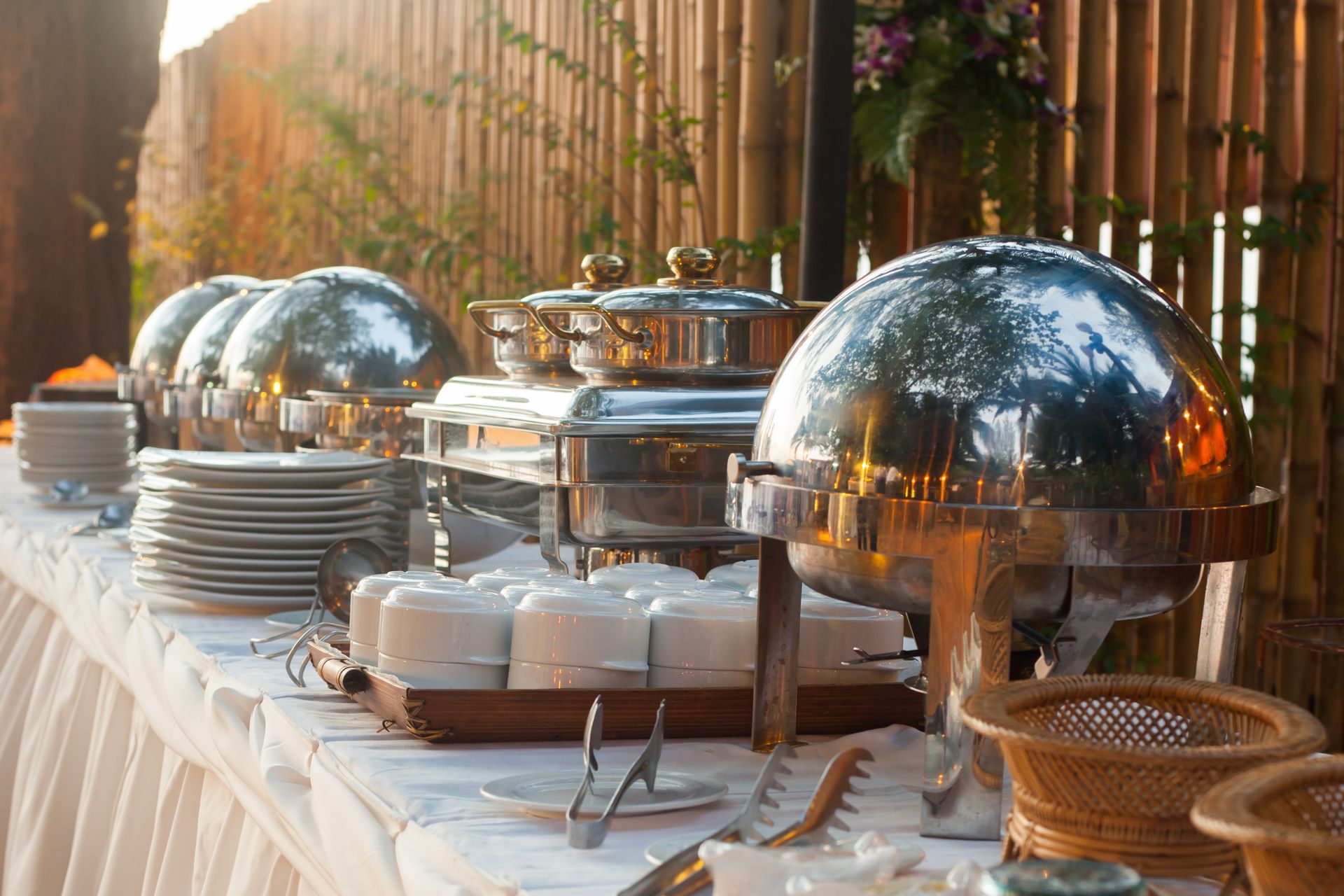 Buffet table with stainless steel chafing dishes and serving utensils, outdoor setting.