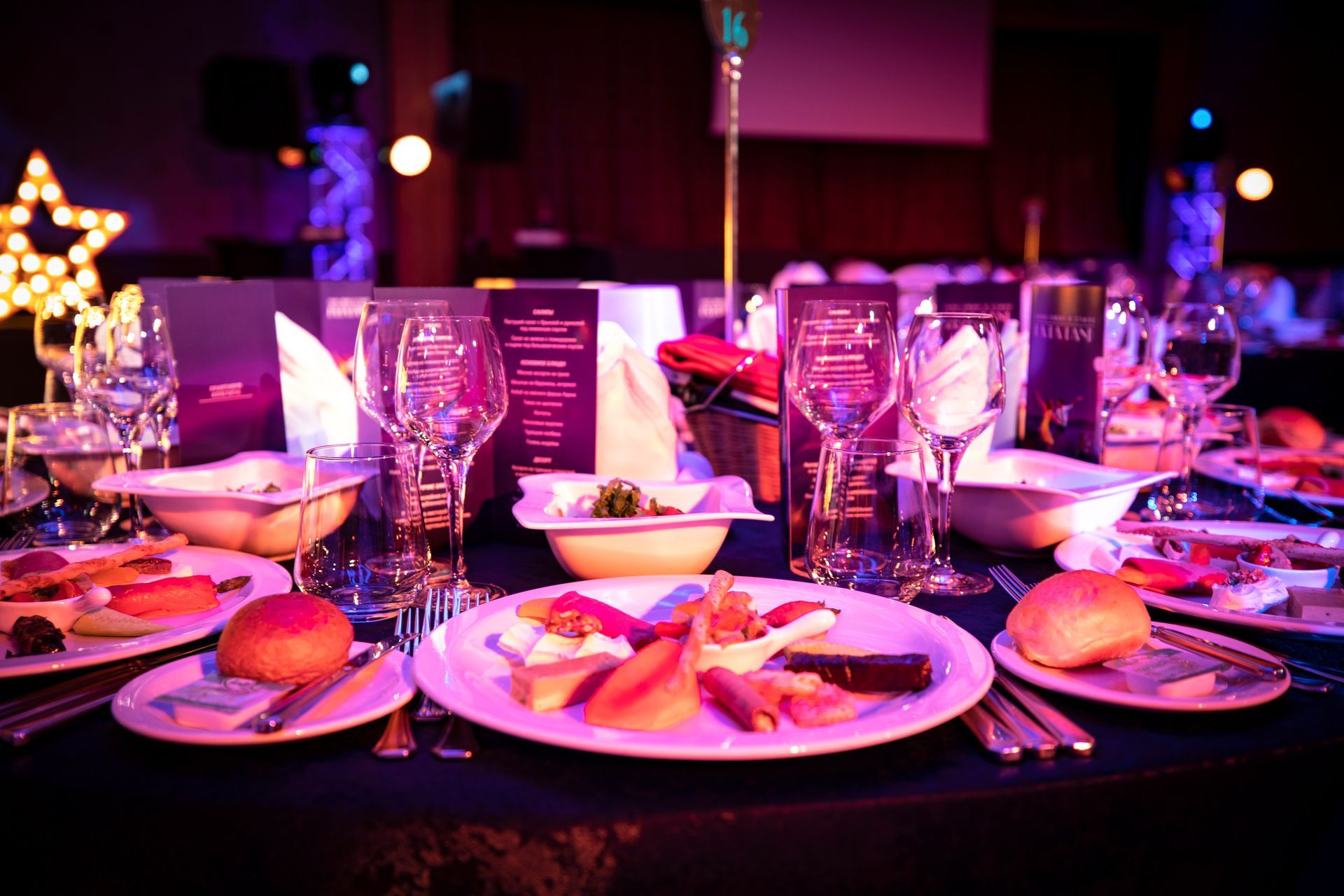 Formal dinner table setting with food, glassware, and menus; venue lit in purple and blue.