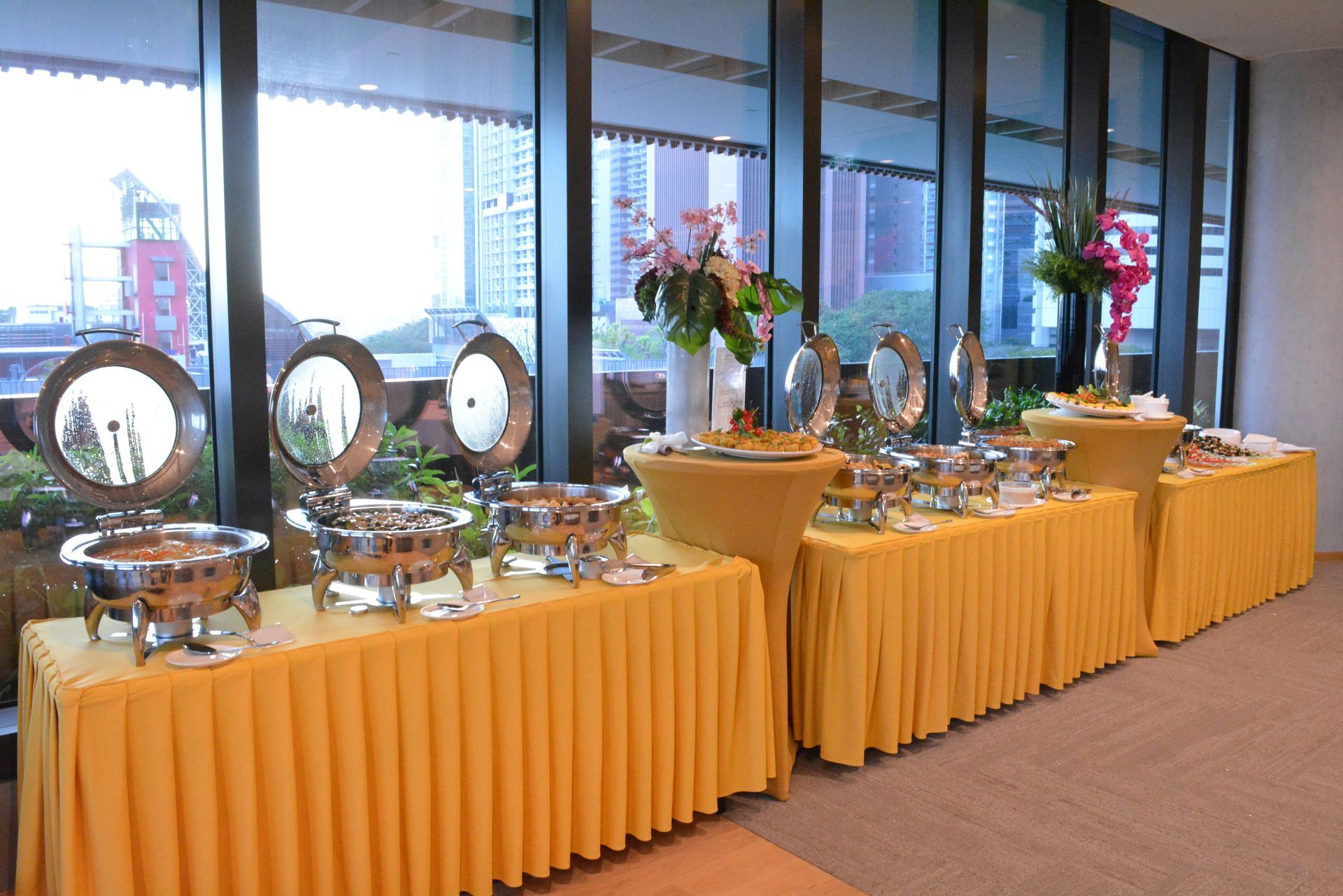 Buffet with golden tablecloths, stainless steel food warmers, flowers, and window view of a cityscape.