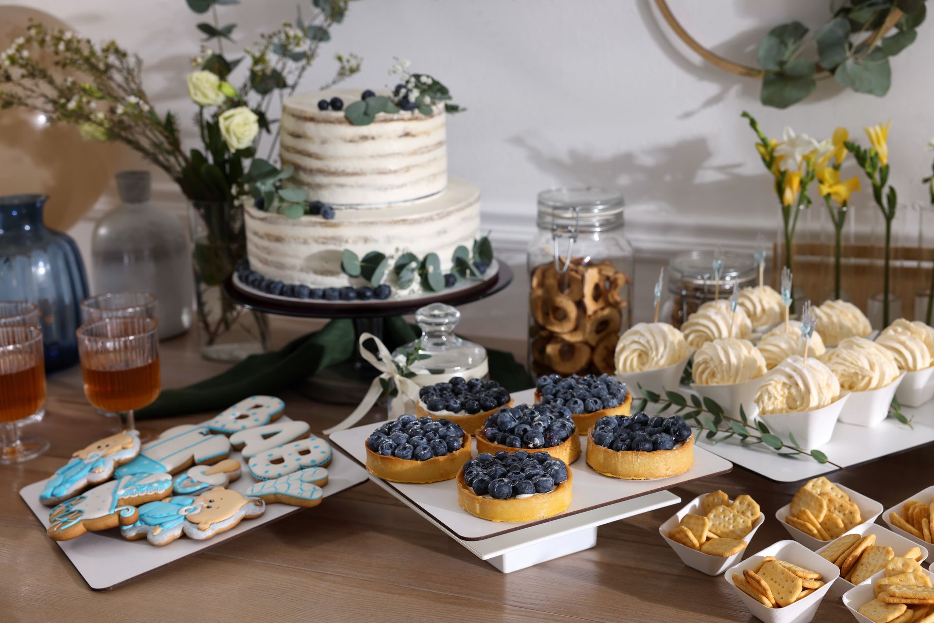 Dessert table: cake, tarts, cookies, and other treats, decorated with blueberries and flowers, for a celebration.