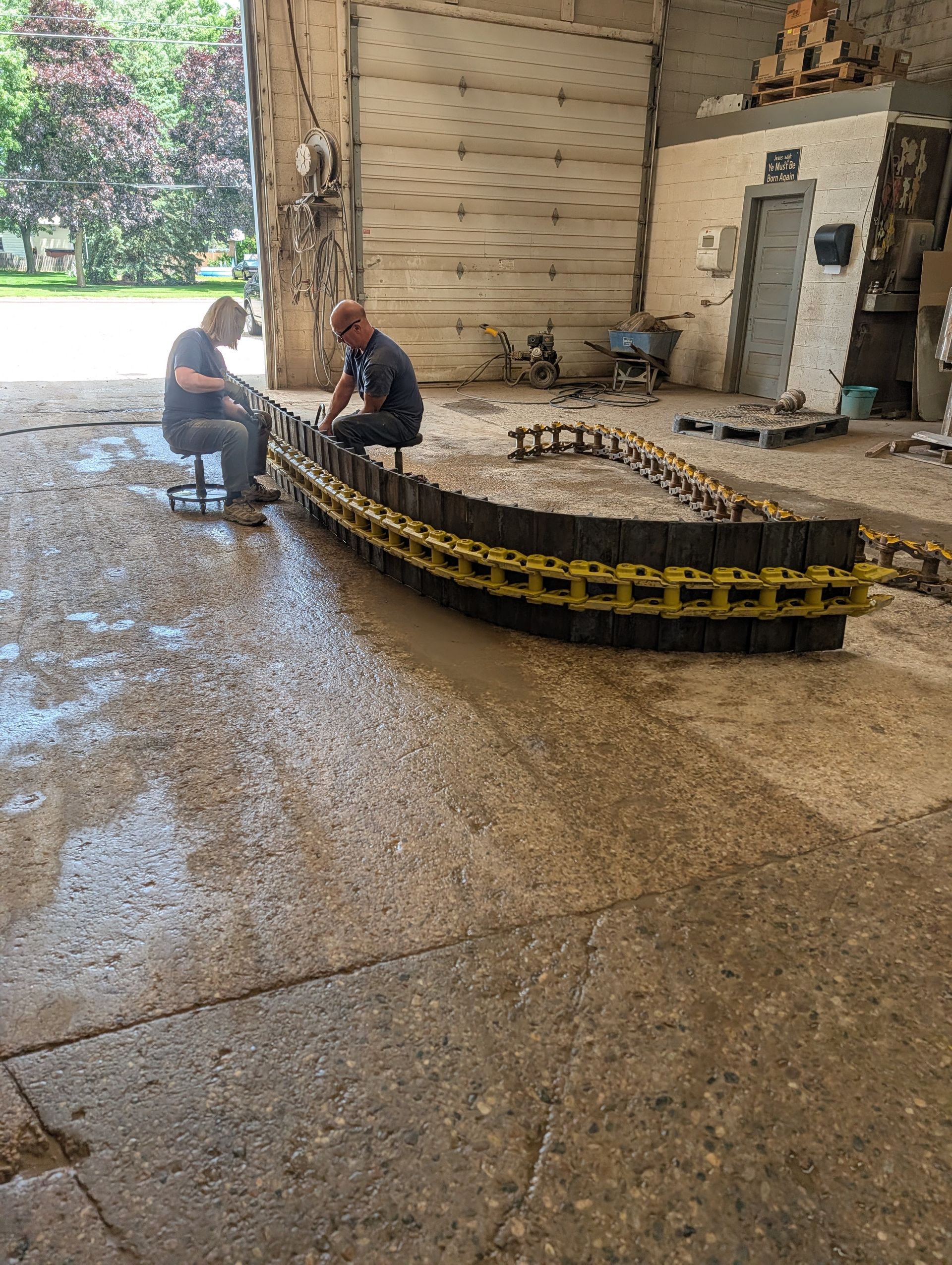 Two workers kneel beside a large metal chain in an industrial workshop.
