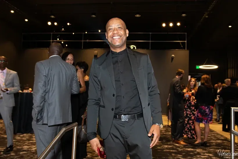 A man in a suit and black shirt is standing in a crowded room.