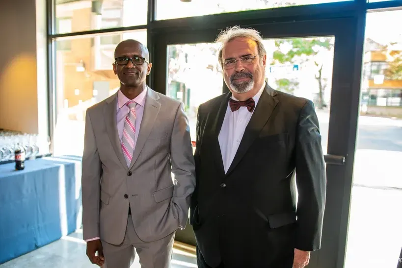 Two men in suits and ties are standing next to each other in front of a window.